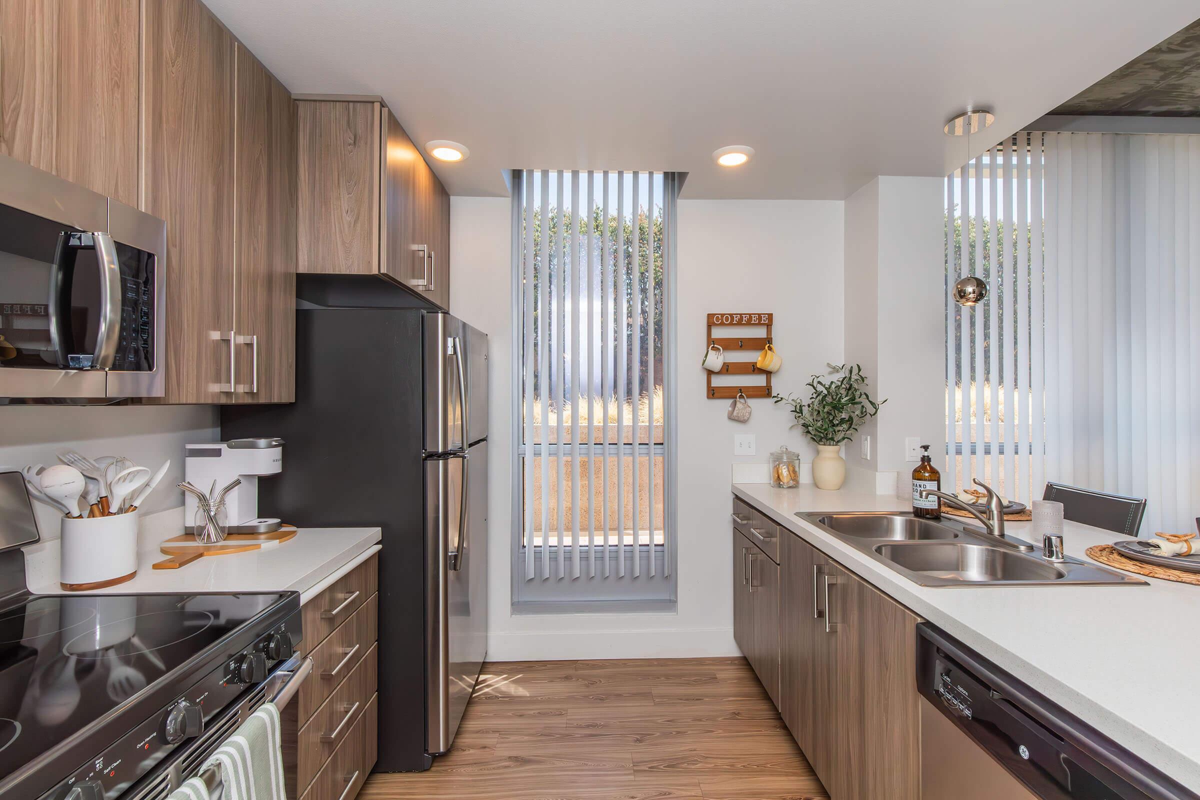Modern kitchen featuring wooden cabinetry, stainless steel appliances, and a white countertop. Includes an inset microwave, stove, and a double sink. Large windows with vertical blinds allow natural light. Decorative items like a plant and a bottle of whiskey enhance the cozy atmosphere.