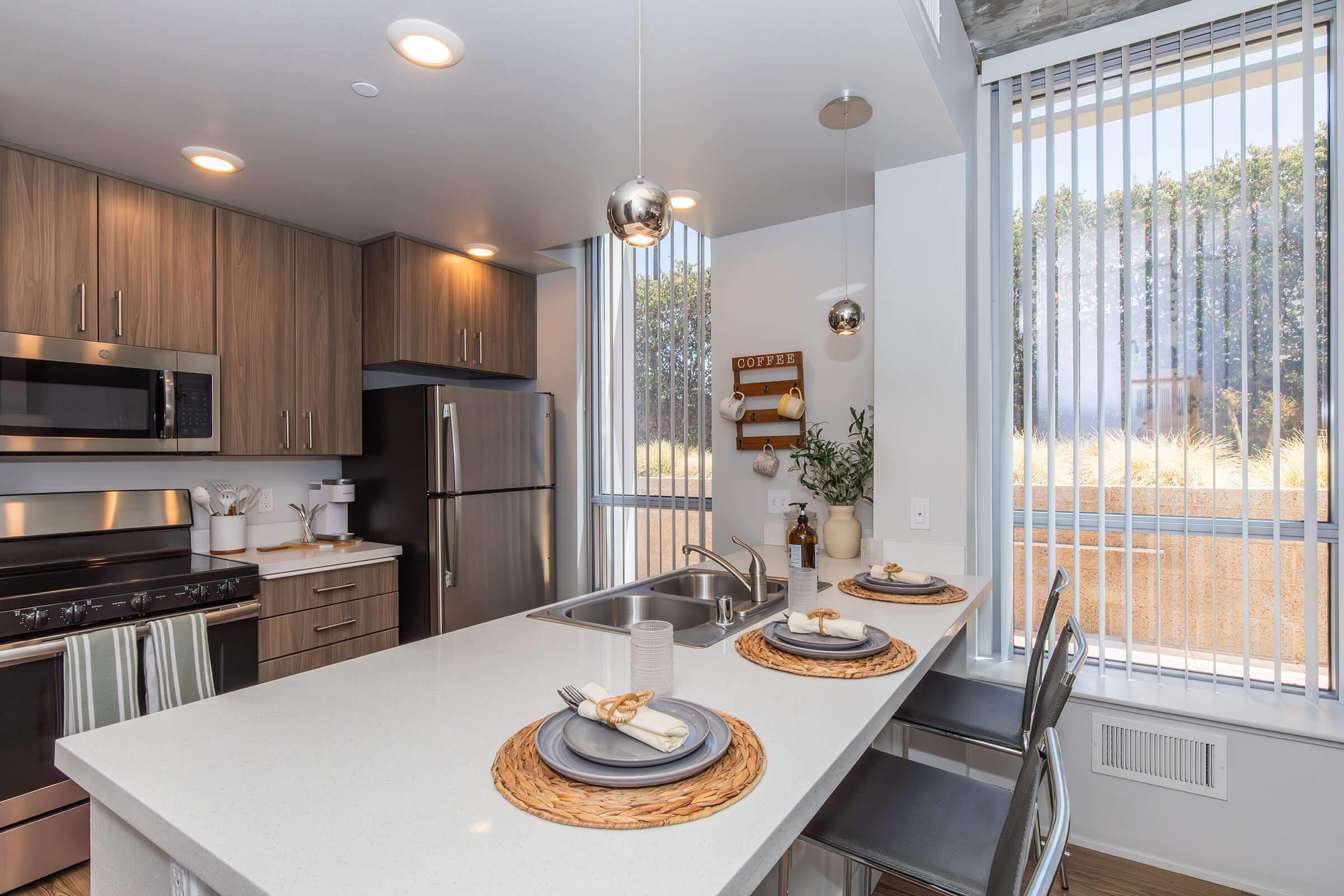 Modern kitchen featuring stainless steel appliances, including a microwave and refrigerator. A spacious island with three place settings, decorative plates, and stylish cutlery. Natural light filters through large blinds, highlighting the warm wooden cabinetry and contemporary decor.