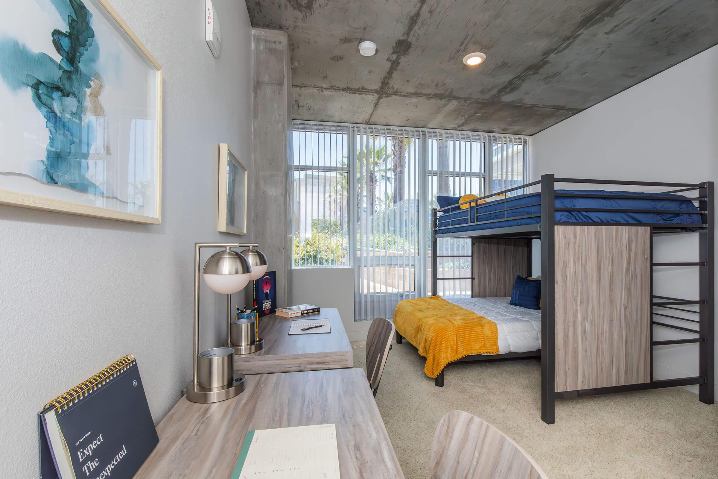 A modern bedroom featuring a lofted bunk bed with blue bedding and a yellow throw, a desk with a lamp and notebook, and a large window with sheer curtains providing natural light. The walls are light-colored with a concrete ceiling, and a plant is visible outside the window.