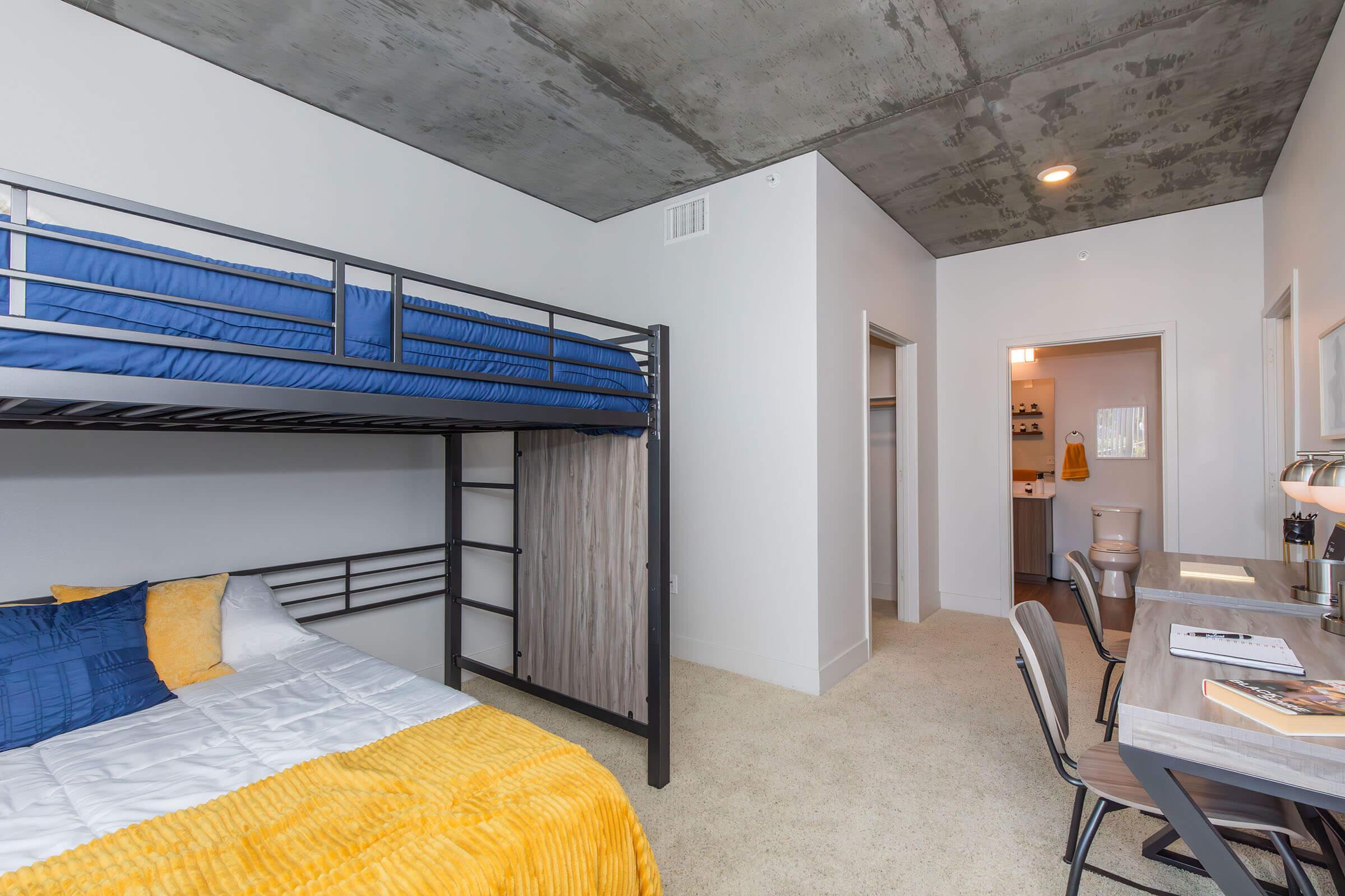 A modern bedroom featuring a lofted bunk bed with a blue top mattress and a yellow-accented lower bed. The room has light-colored walls, a concrete ceiling, and a desk with a chair. A doorway leads to another room, visible in the background, which includes a small kitchen area and bathroom.