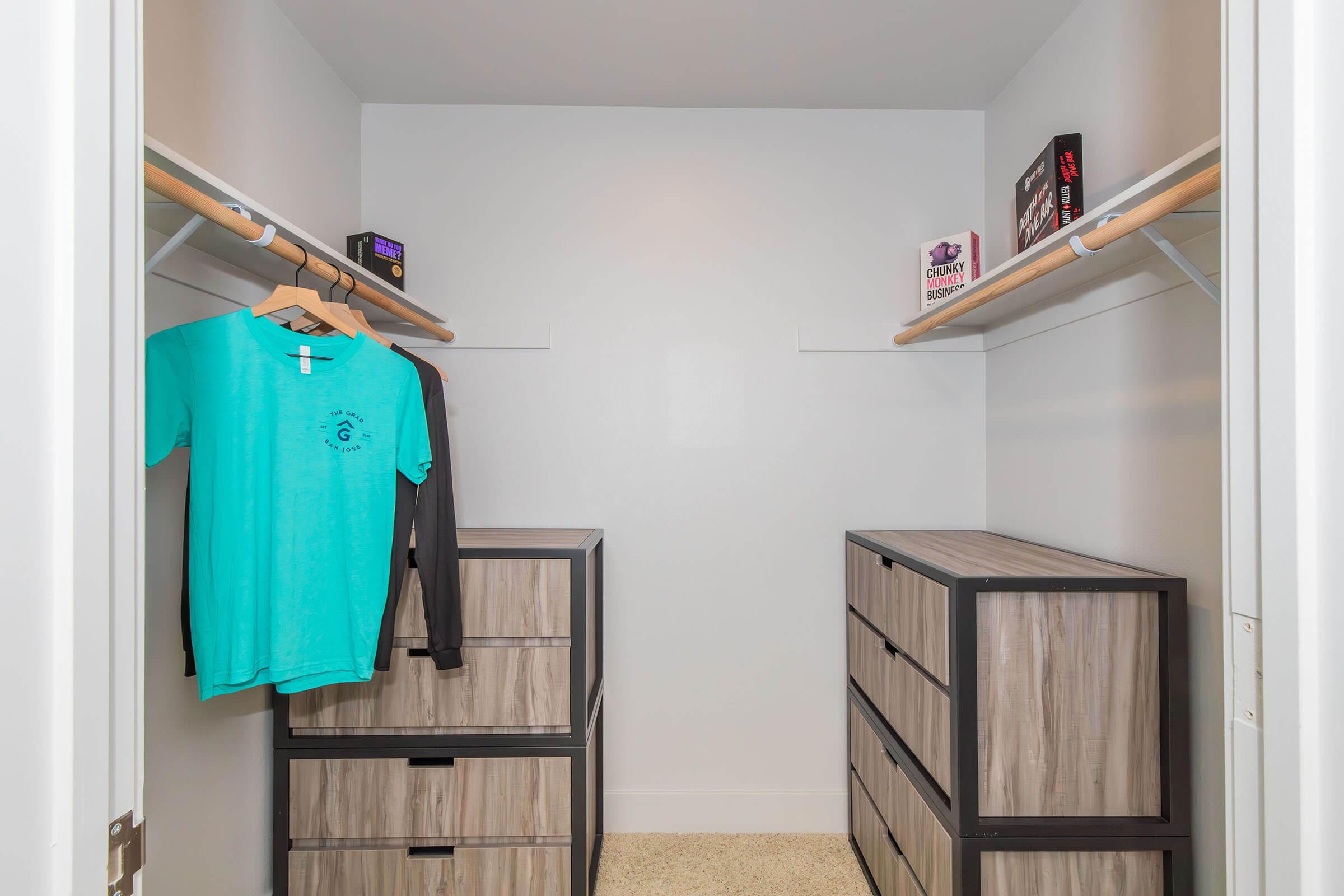 A well-organized closet featuring two clothes hanging rods, a turquoise t-shirt, and a long-sleeved shirt on hangers. There are two modern storage drawers in the center, and boxes on top of them. The walls are painted white, creating a bright and clean appearance.