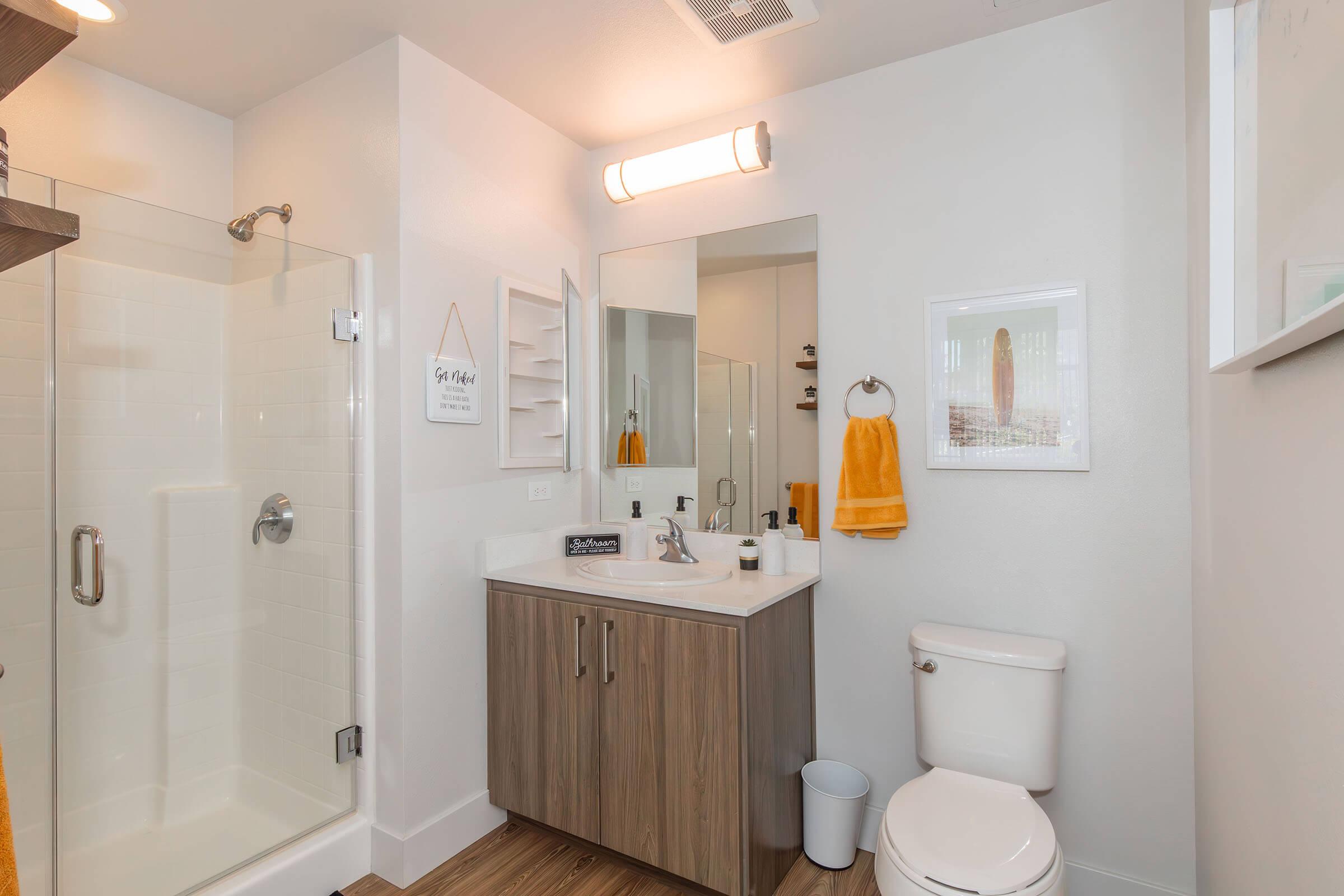 Modern bathroom featuring a glass shower enclosure, a double-sink vanity with a light wooden finish, a mirror, and a towel rack. The decor includes orange towels, a small decorative sign, and a white closet shelf. Natural light from a window highlights the clean, minimalist design.