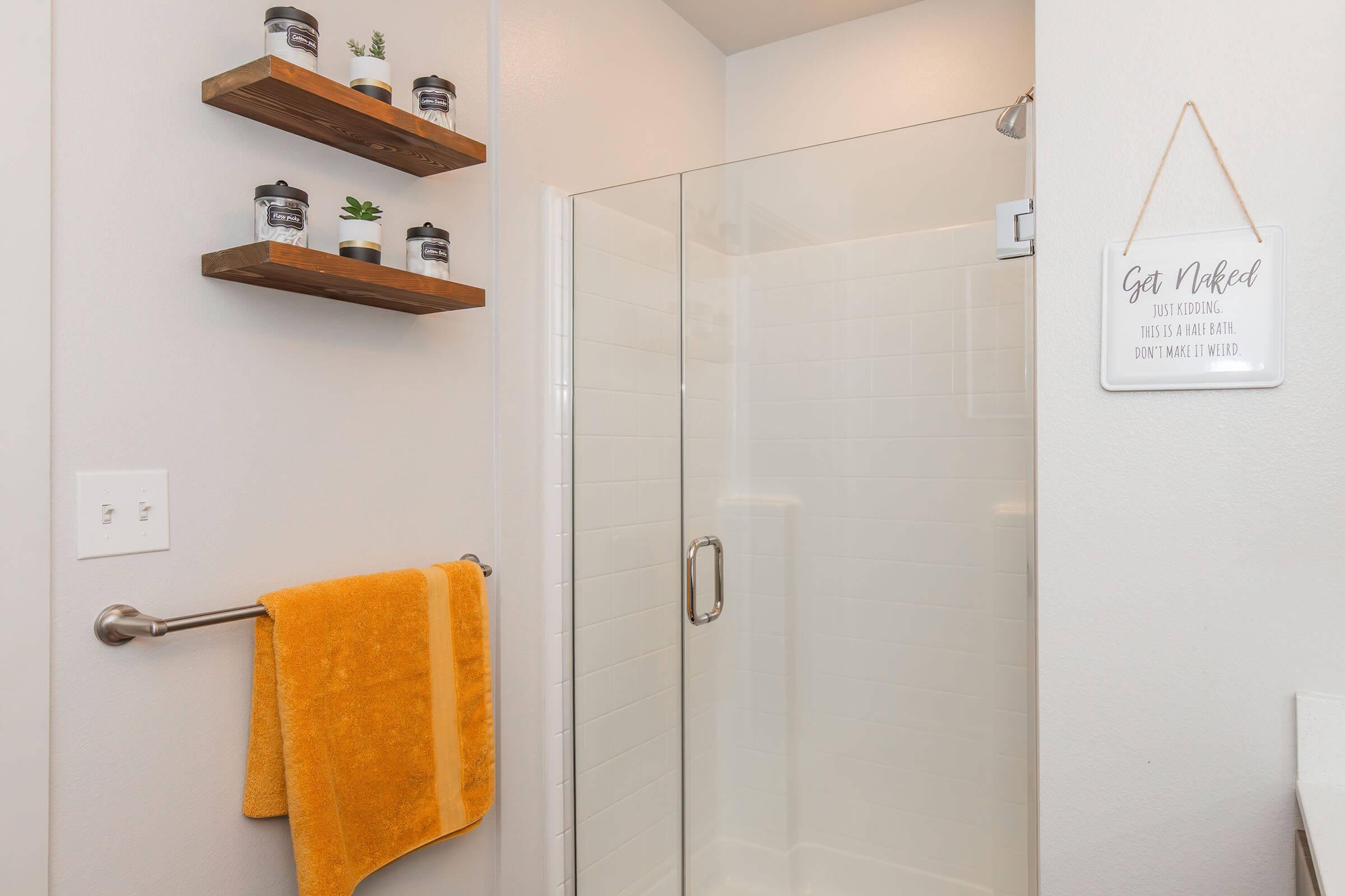 A modern bathroom featuring a glass shower enclosure, wall shelves adorned with small potted plants, a yellow towel hanging on a towel bar, and a decorative sign that reads "Get Naked. Just kidding! This is a bathroom. Don't make it weird." The walls are painted a light color, contributing to a clean aesthetic.