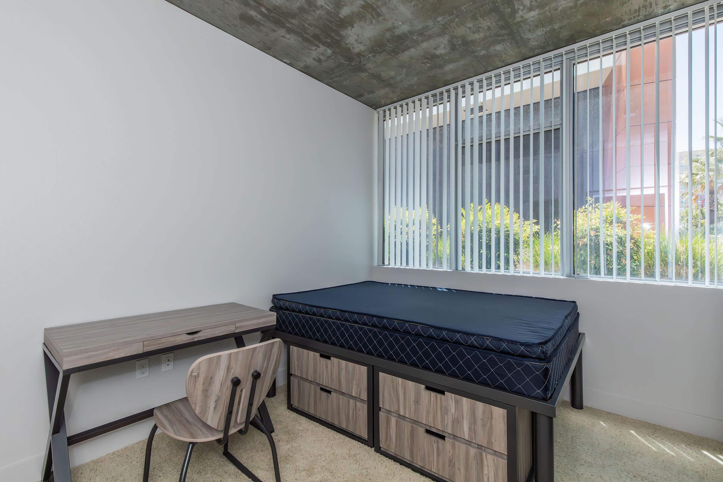 A minimalist bedroom featuring a bed with a blue mattress, a wooden desk with a chair, and large windows with blinds allowing natural light. The room has a concrete ceiling and light-colored walls, with a carpeted floor. Outside the window, greenery is visible.