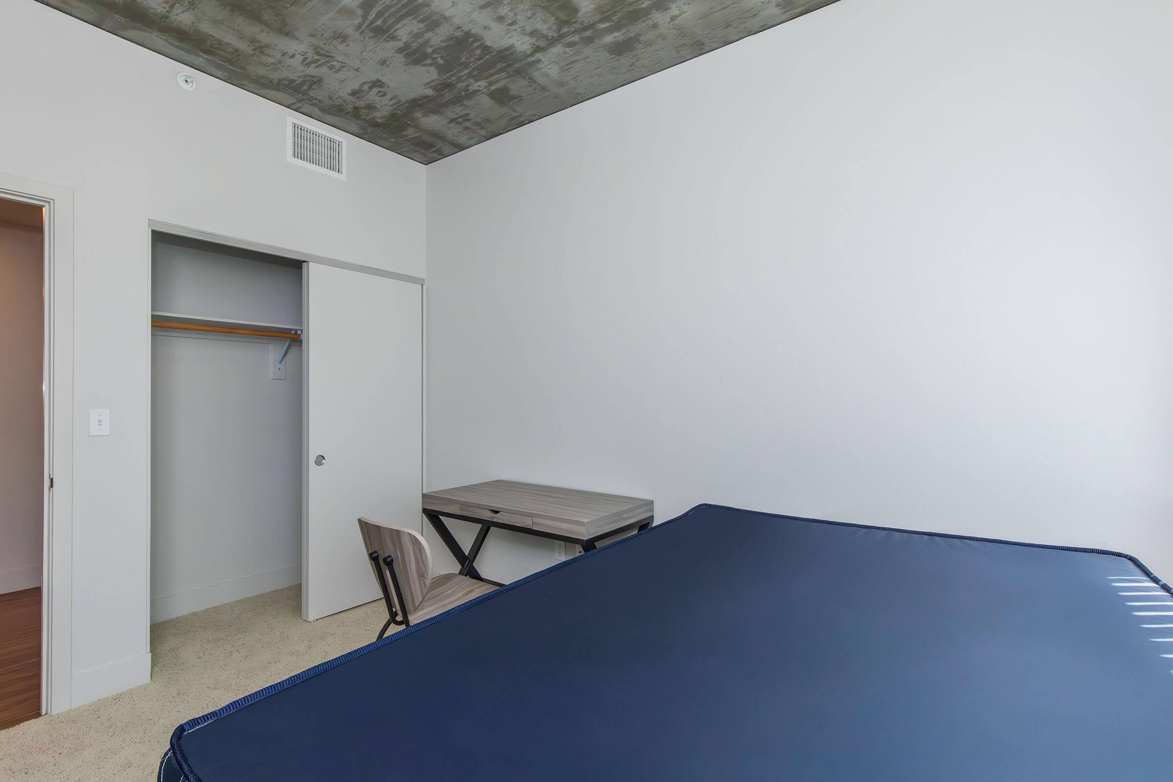 A minimalist bedroom featuring a blue mattress on a bed frame, a small wooden desk with a chair, and an open closet with a hanger. The walls are painted white, and the ceiling has a concrete finish, creating a modern and clean aesthetic.