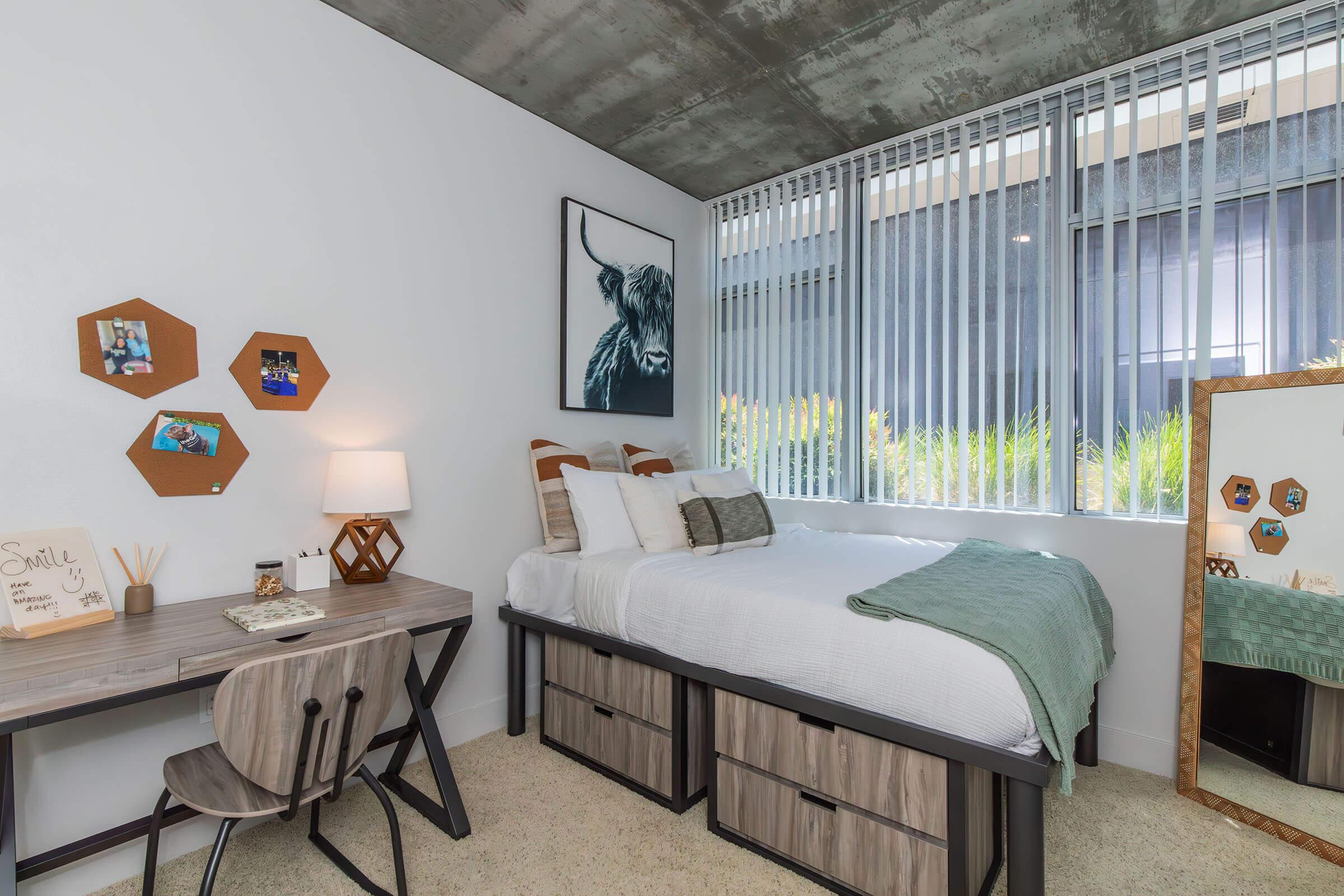 A modern bedroom featuring a queen-sized bed with decorative pillows, a green throw blanket, and a large window with vertical blinds. A wooden desk with a chair is placed next to the bed, adorned with a lamp and a framed picture. Honeycomb shelves display photos, and a mirror reflects the room's furnishings.