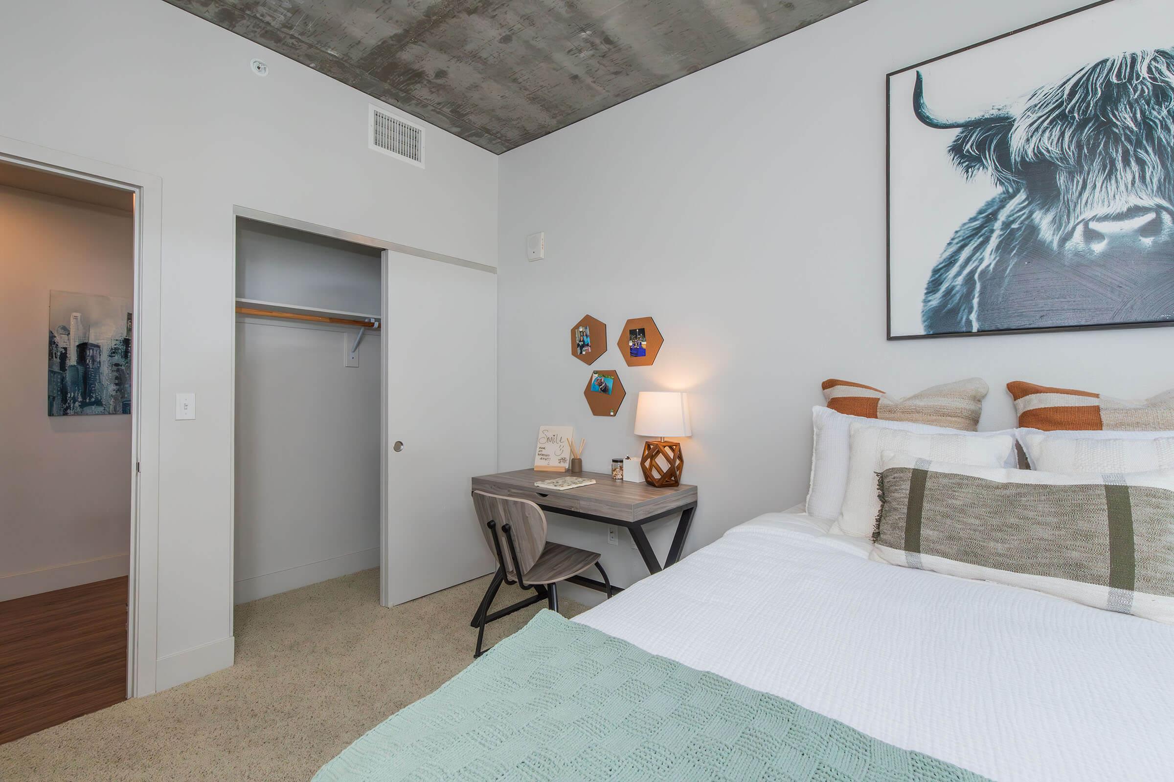 A cozy bedroom featuring a queen-sized bed with decorative pillows, a small desk and chair, a closet, and a wall art of a cow. The room has a modern design with concrete ceiling, neutral walls, and carpeted flooring, creating a warm and inviting atmosphere.