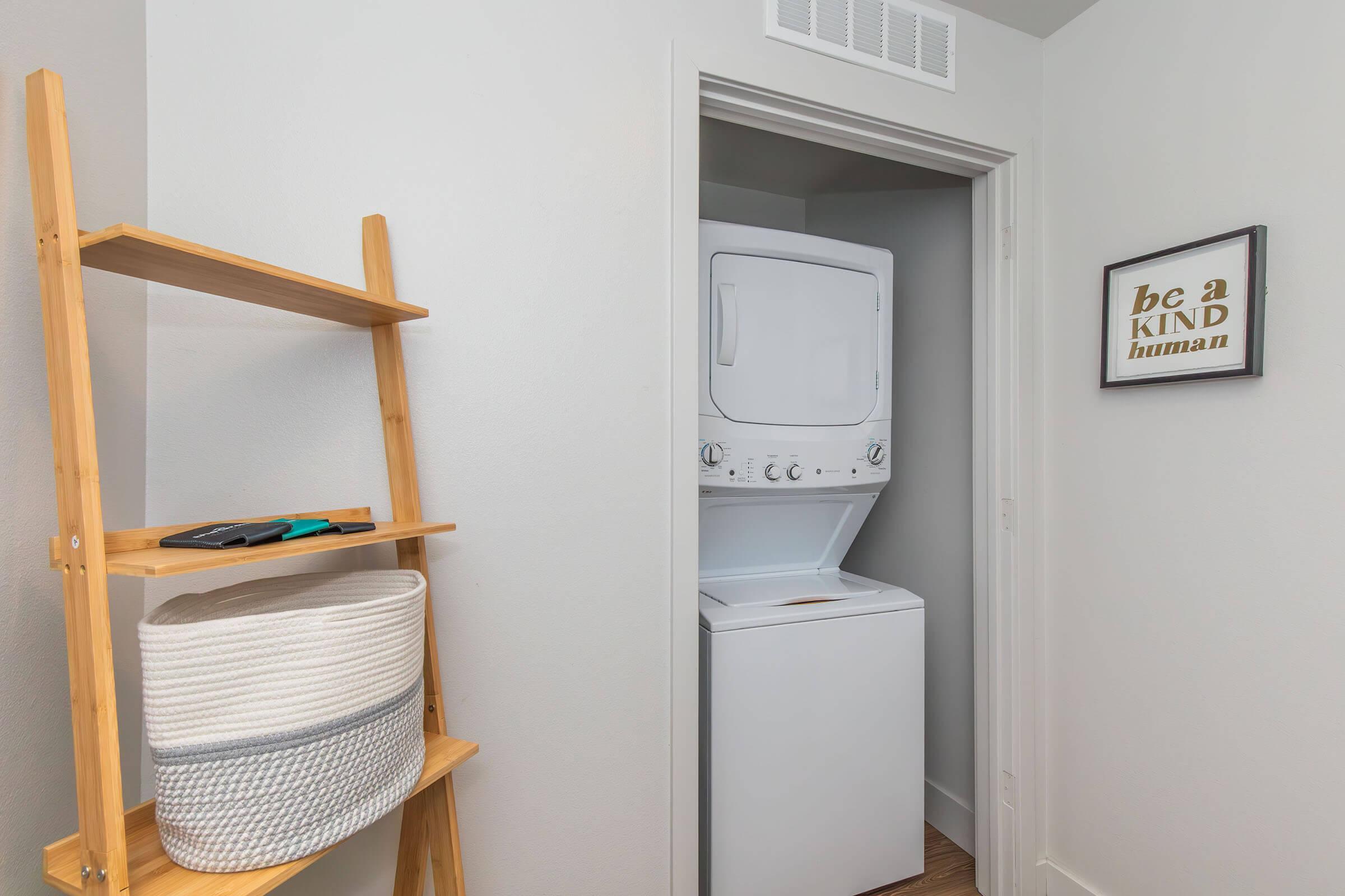 A small laundry area featuring a stacked washer and dryer in a closet, complemented by a wooden shelf holding a basket and some accessories. On the wall, there's a framed artwork with the text "be a kind human" adding a personal touch to the space. The decor is modern and minimalistic.