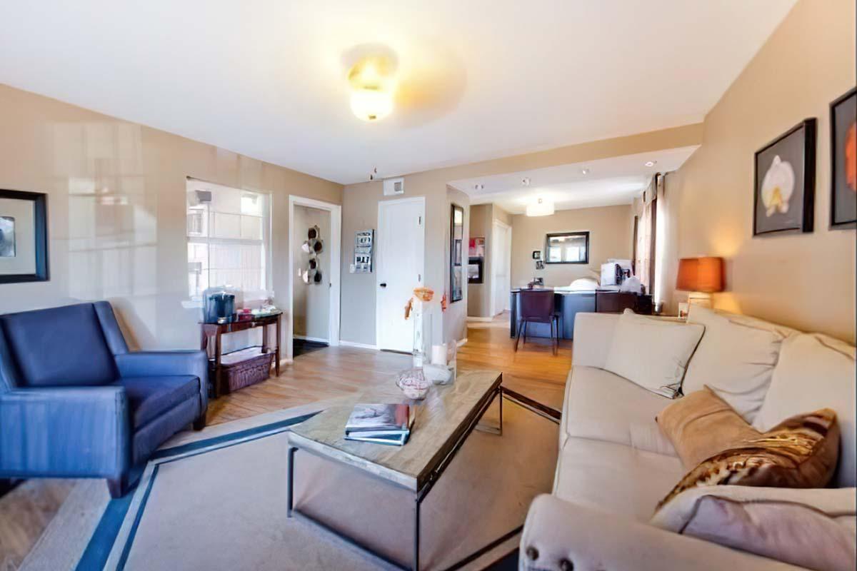 A cozy living room featuring a beige sofa and a blue armchair, a glass coffee table on a light rug, and decorative art on the walls. The space has warm lighting, wooden flooring, and leads to a dining area in the background, creating an inviting atmosphere for relaxation and socializing.