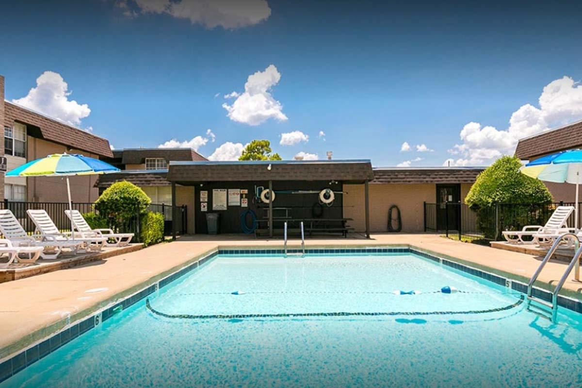 A clear swimming pool surrounded by lounge chairs under colorful umbrellas, with a building in the background. The scene is set against a bright blue sky with a few clouds, creating a relaxing outdoor atmosphere.