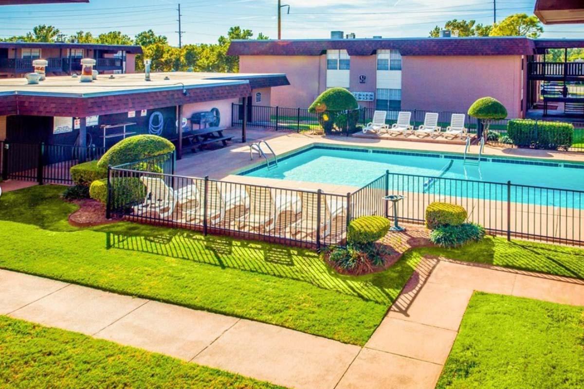 A bright outdoor scene featuring a swimming pool surrounded by neatly arranged lounge chairs. Lush green grass and well-trimmed bushes create a welcoming atmosphere. Sunlight casts a warm glow over the area, with nearby buildings providing a cozy backdrop. The setting is ideal for relaxation and leisure.