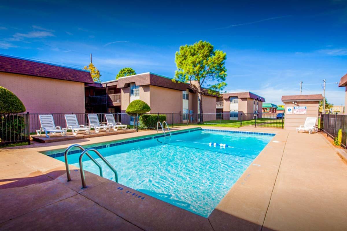 A tranquil outdoor swimming pool surrounded by well-maintained landscaping and lounge chairs. The pool is clear and inviting, with nearby apartment buildings and a bright blue sky overhead. Fencing encloses the area, enhancing the privacy of this relaxing space.