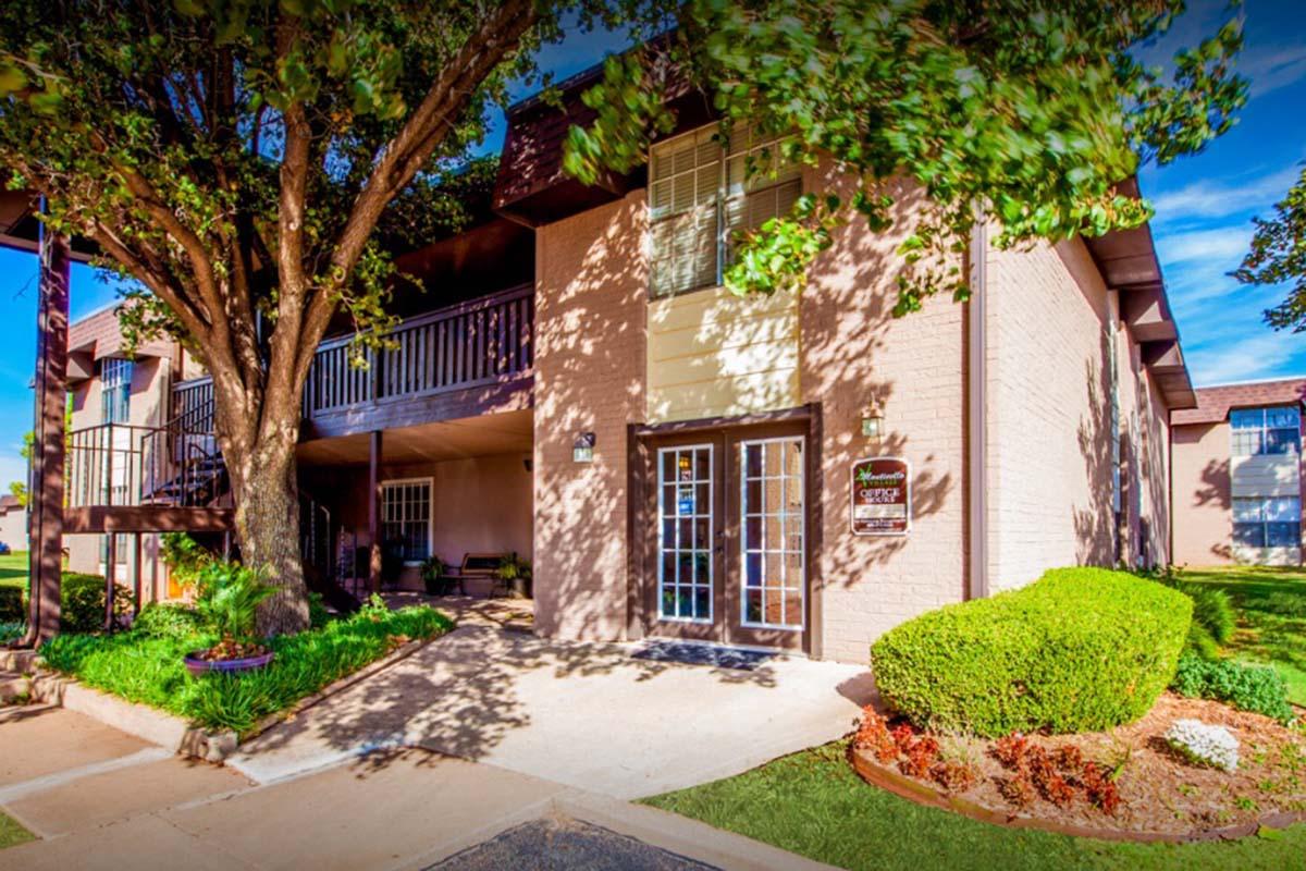 A well-maintained two-story building with a light-colored exterior, surrounded by lush green landscaping and a large tree. The entrance features double doors and a small porch area, with a staircase leading to the upper level. The setting is bright and inviting, typical of an apartment complex.
