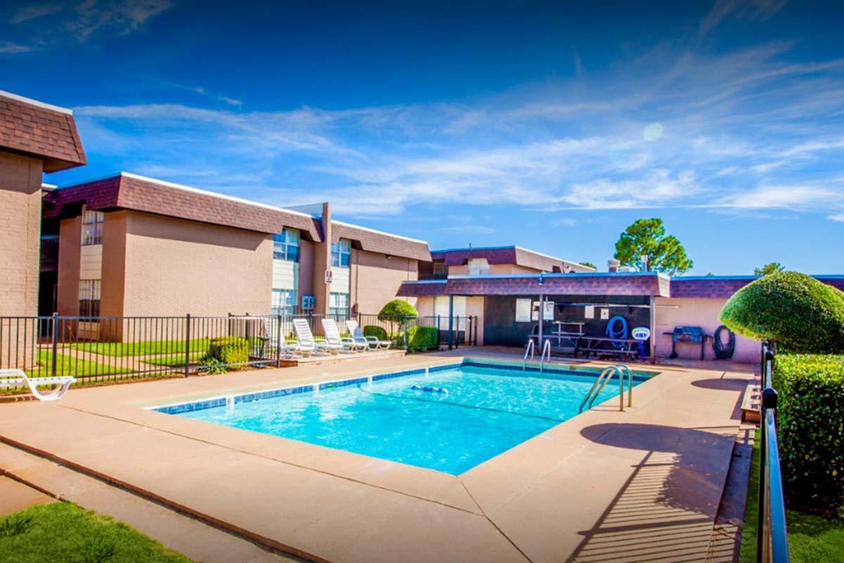 A sunny outdoor swimming pool surrounded by a fence, with lounge chairs nearby. Adjacent to the pool is a small building with blue accents, and well-maintained landscaping featuring neatly trimmed bushes. The sky is clear with a few clouds, creating a vibrant and inviting atmosphere.