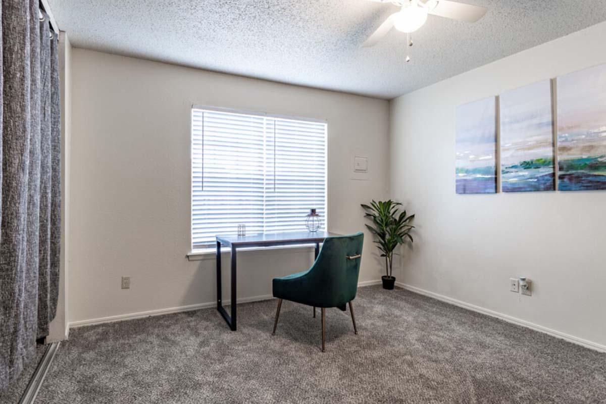 A cozy room featuring a small desk with a plant, a green chair, and three abstract paintings on the wall. There is a large window with blinds allowing natural light in, and the floor is covered with gray carpet. Light-colored walls create a bright and inviting atmosphere.