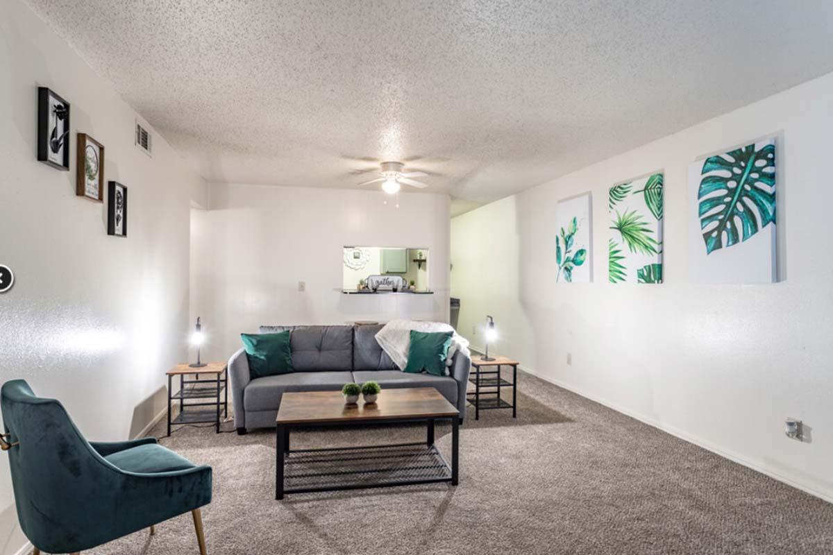 A cozy living room featuring a grey sofa with teal cushions, a wooden coffee table with small plants, and two stylish side tables with lamps. The wall displays framed art and three canvas prints of green leaves, while the carpeted floor adds warmth to the space. A small kitchenette is visible in the background.