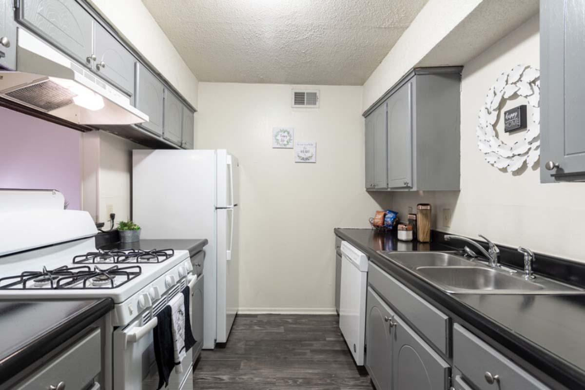 A modern kitchen featuring gray cabinets, a white stove and refrigerator, a dishwasher, and a double sink. The countertops are dark, and there are decorative elements on the walls including a round mirror and framed pictures. The flooring is dark wood.
