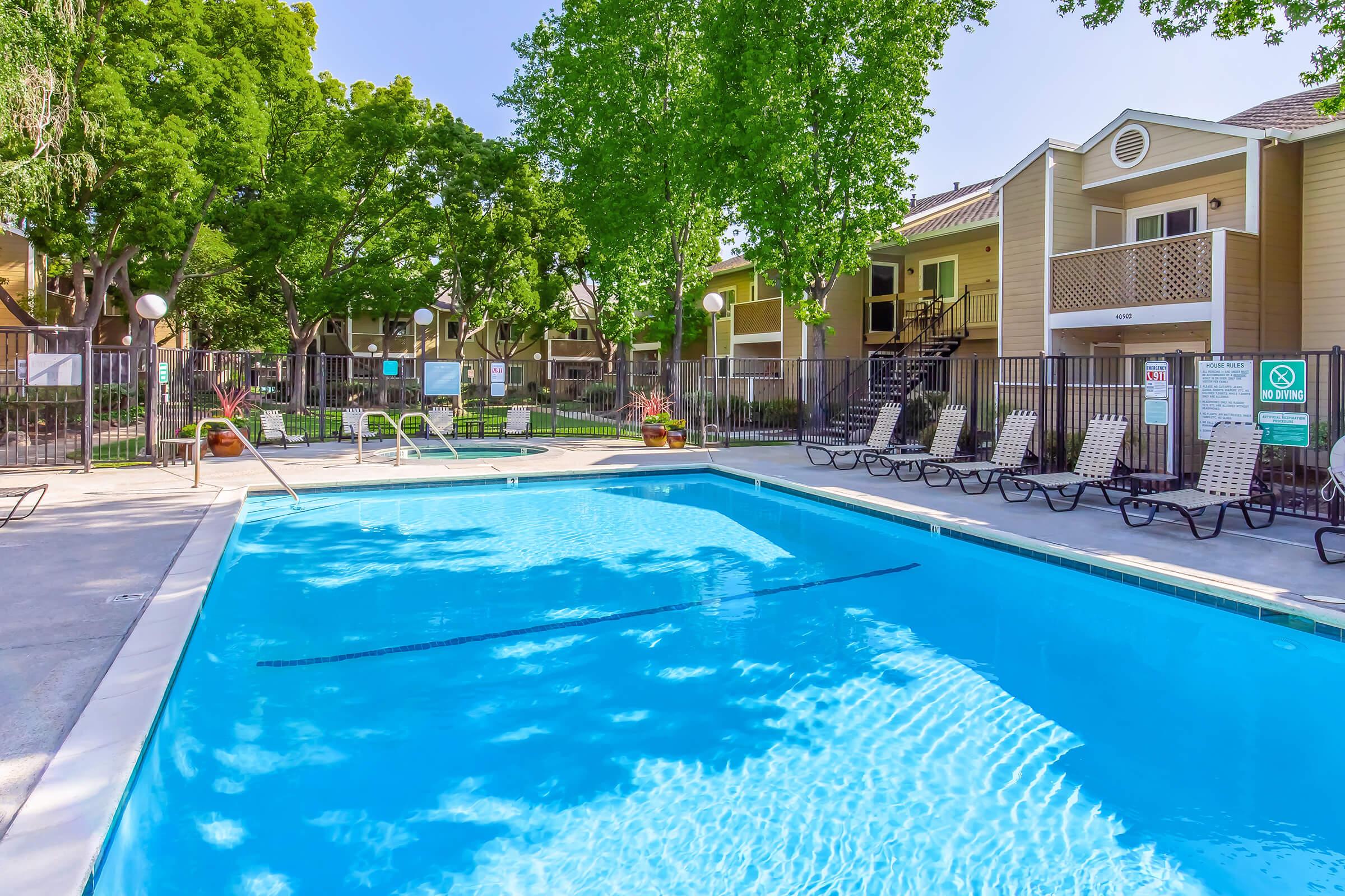 A vibrant swimming pool surrounded by lounge chairs, located in a well-maintained outdoor area with trees and apartment buildings in the background. The clear blue water reflects the sunny sky, creating a welcoming atmosphere for residents and guests.