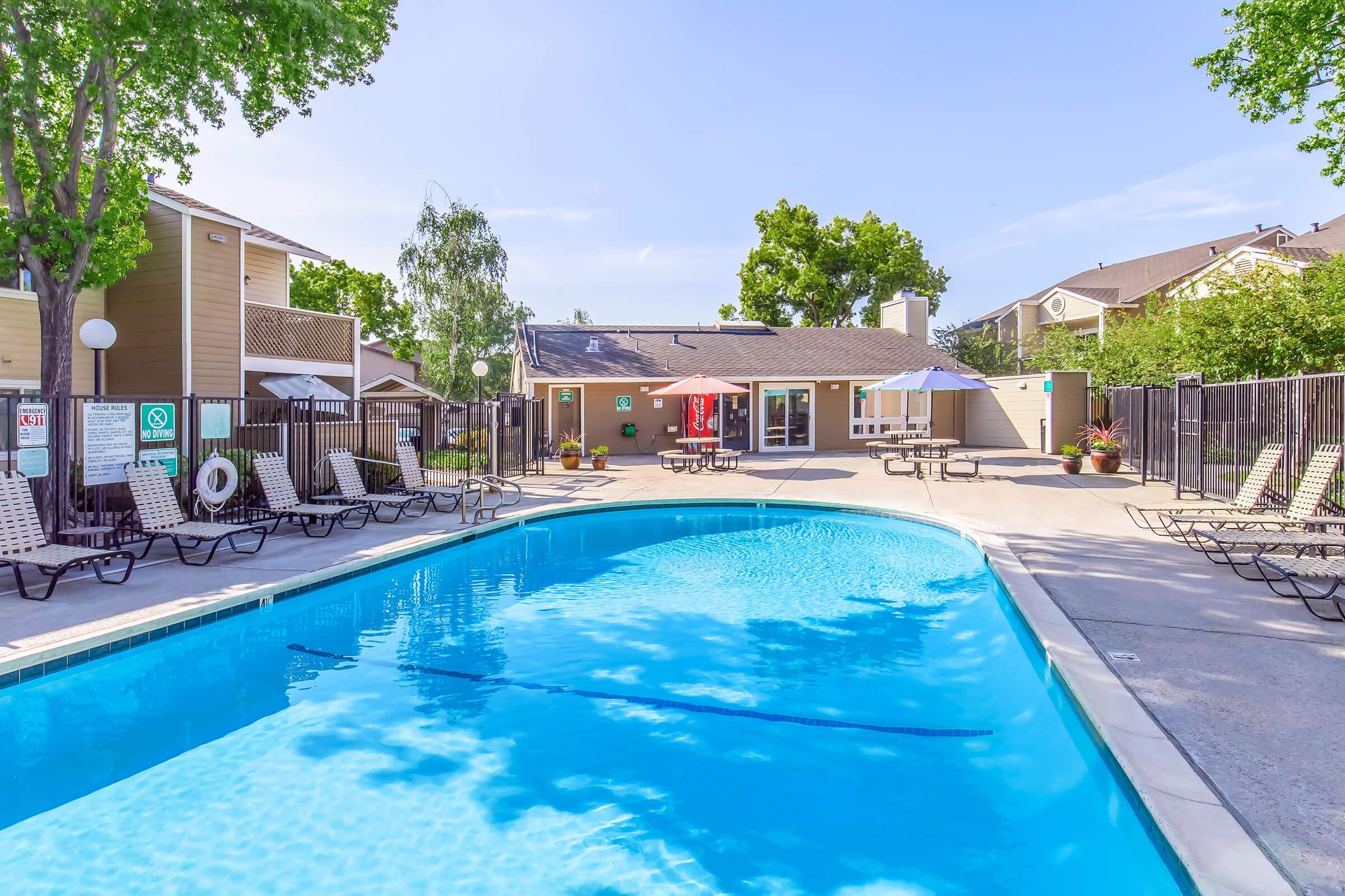 A clean swimming pool surrounded by lounge chairs and umbrellas, with a clubhouse visible in the background. Lush trees and blue skies add to the inviting atmosphere of this outdoor leisure area.
