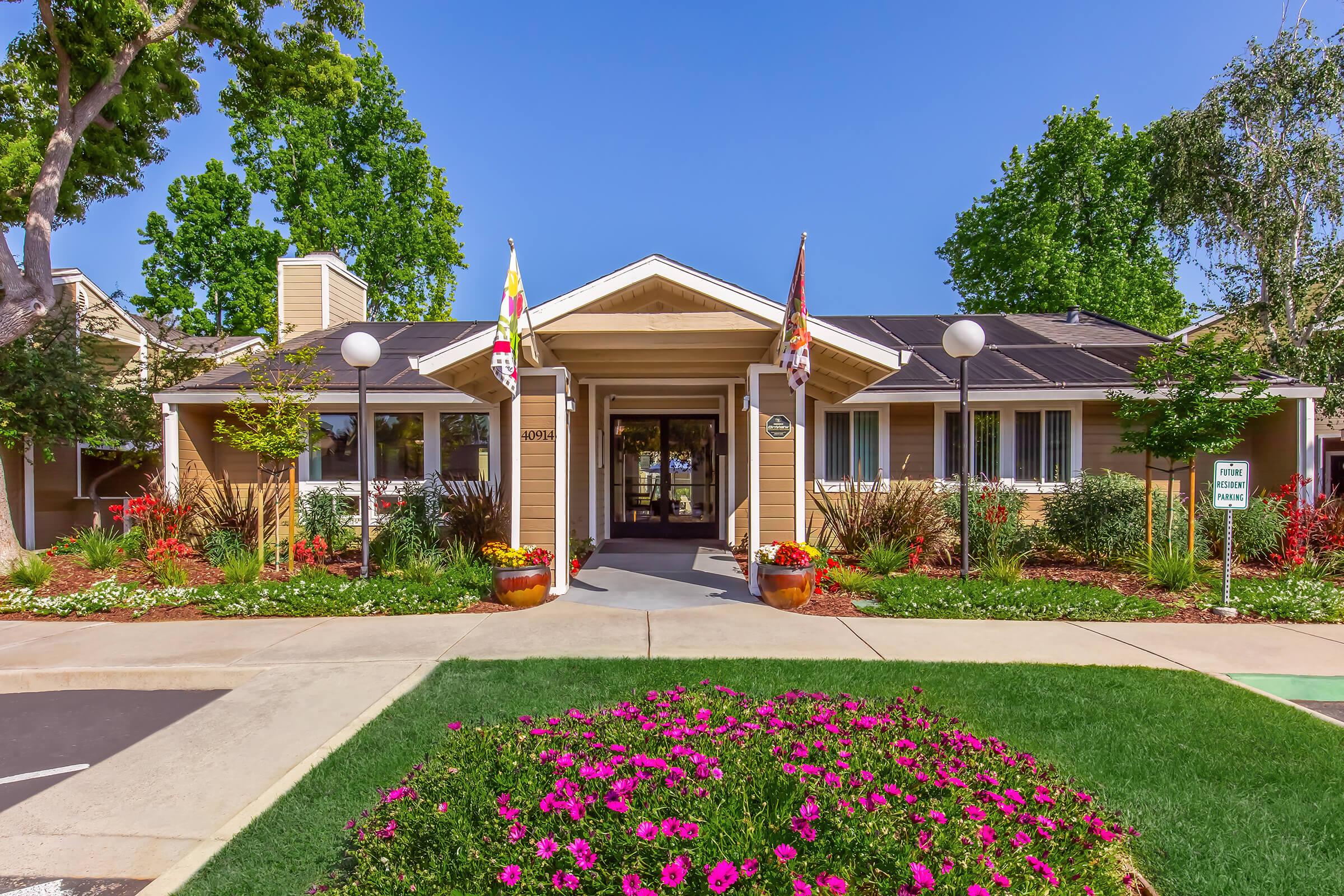 A charming building with a welcoming entrance, featuring large front windows, colorful flower beds, and landscaped greenery. Flags are displayed outside, and the pathway leads to the main door, enhancing the inviting atmosphere of the property. Clear blue sky overhead adds to the cheerful scene.