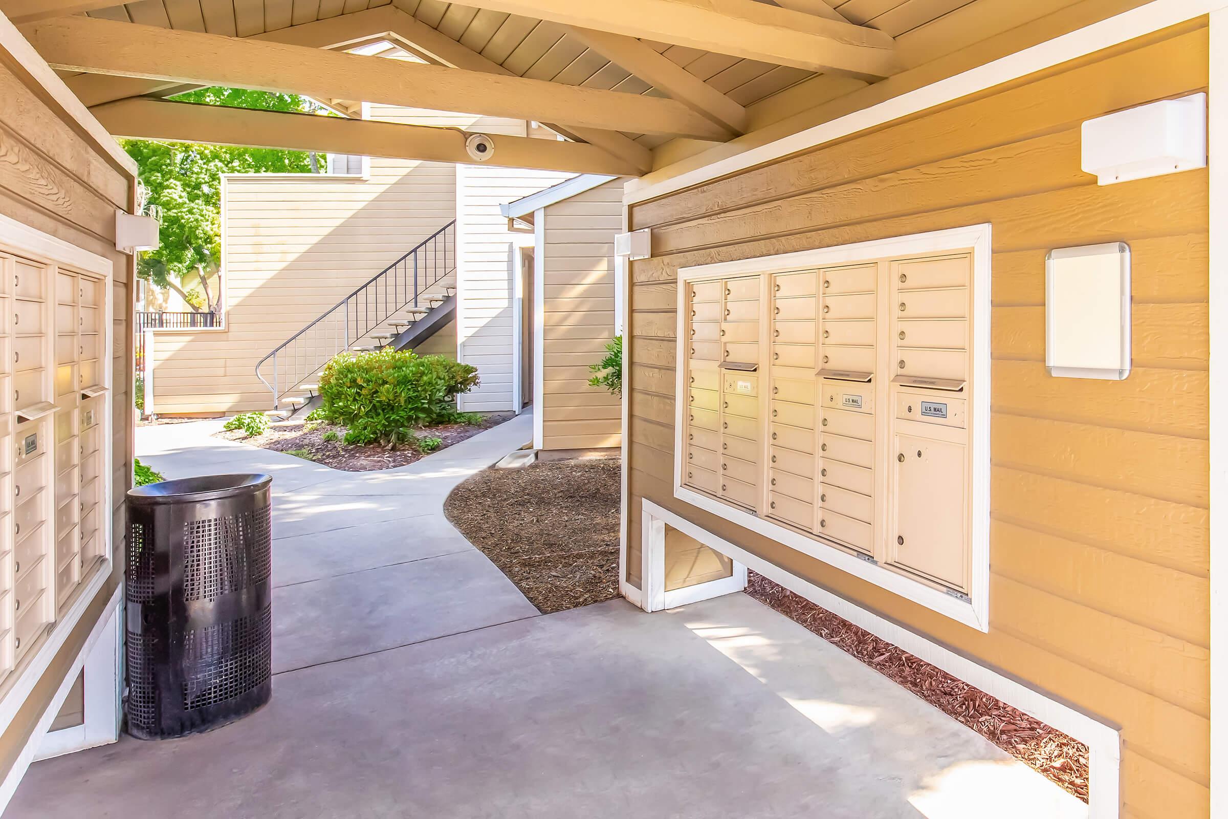 A walkway in an apartment complex featuring mailboxes mounted on the wall, a black trash can, and a landscaped area with bushes. In the background, outdoor stairs lead to additional residential units. The setting is bright and well-maintained, with a focus on convenience and accessibility.