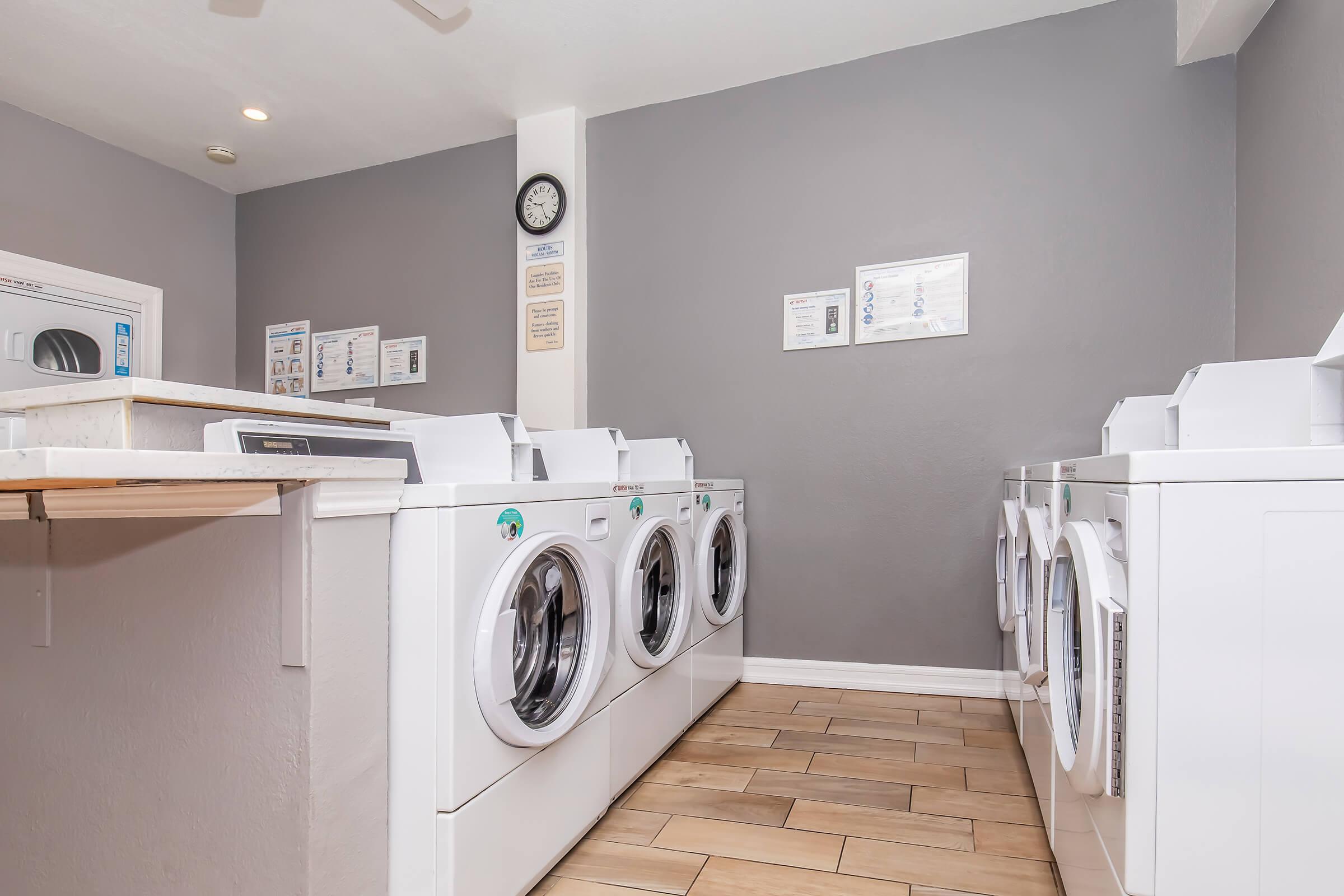 Interior of a modern laundry room featuring multiple white washing machines and dryers lined up against a gray wall. A clock is visible on the wall, along with informational signs. The flooring is tiled in a light color, creating a clean and functional environment.