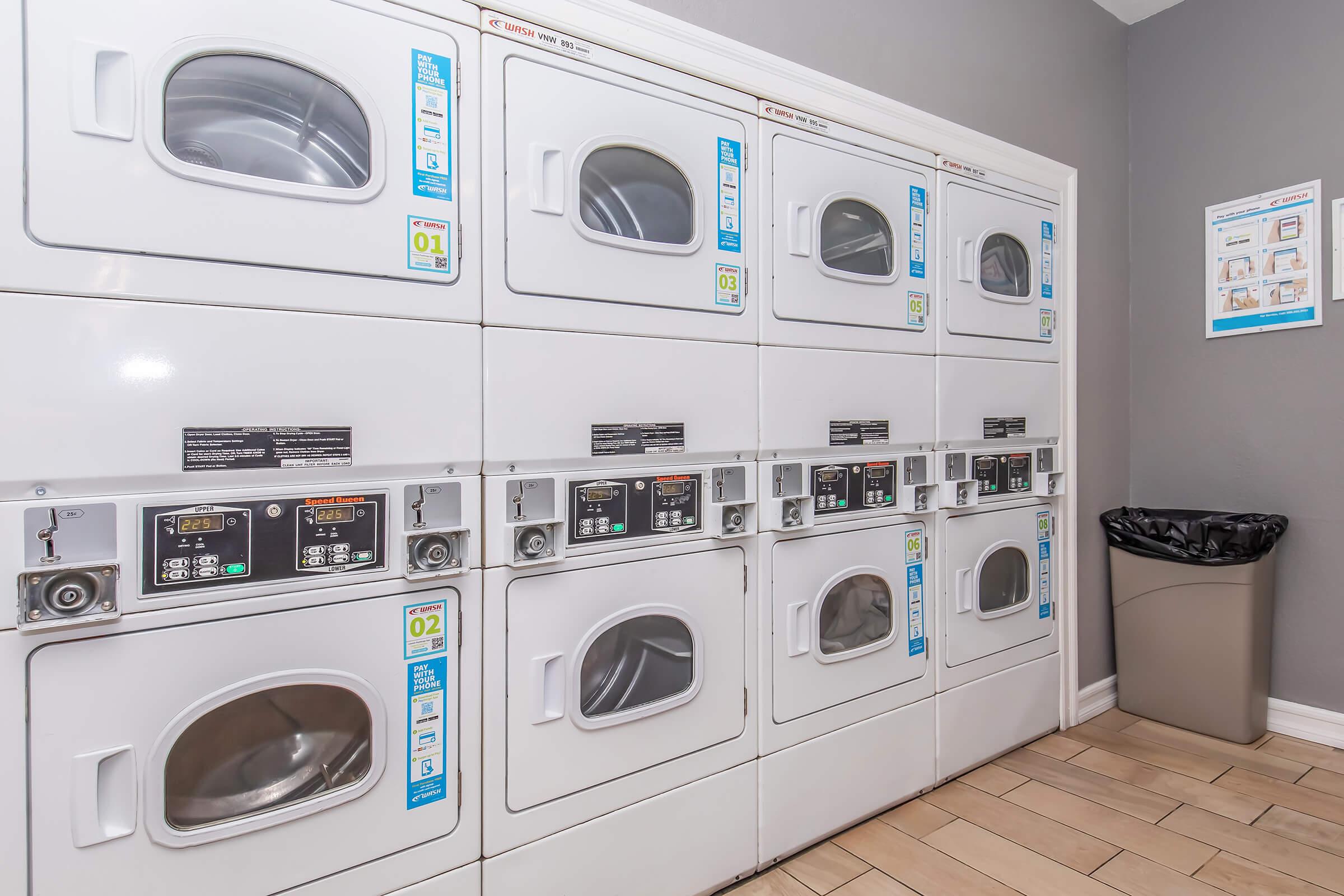 A clean laundry room featuring a row of modern, stacked dryers with digital control panels. Each dryer has clear labels for operation and settings. There is a trash bin in the corner and a wall poster with laundry instructions. The walls are painted a neutral gray, adding a bright, organized feel to the space.