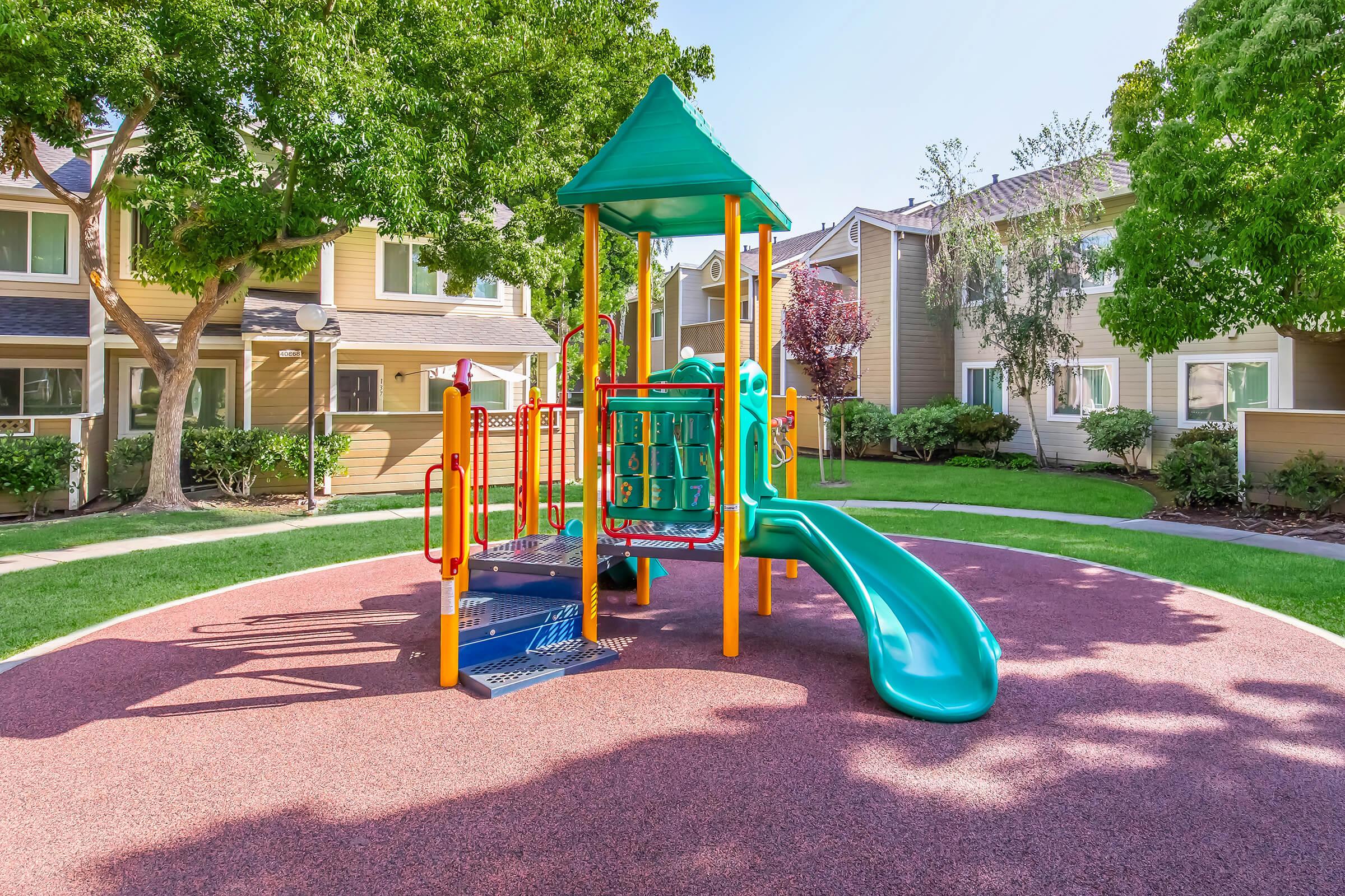 A colorful playground with a slide and climbing structures, situated on a soft rubber surface. The area is surrounded by green grass and trees, with residential buildings visible in the background. The sunny day adds to the cheerful atmosphere.