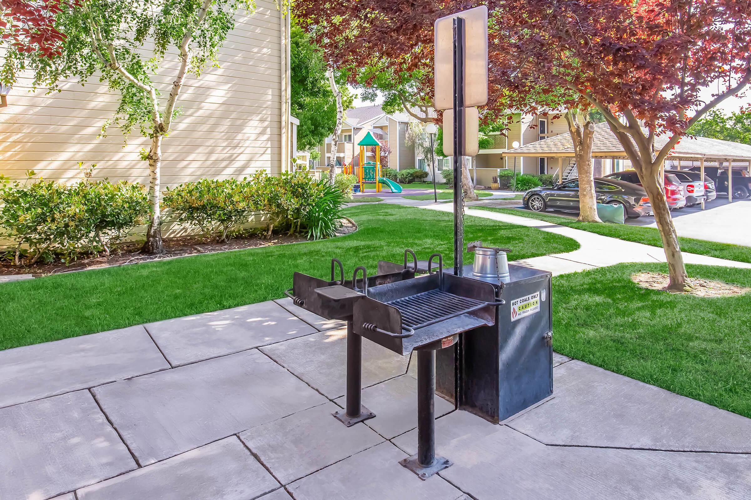 Outdoor barbecue grill in a residential area, situated on a concrete slab with green grass around it. A stop sign is visible nearby, along with trees and a playground in the background. The scene includes parked cars and a well-maintained landscape.