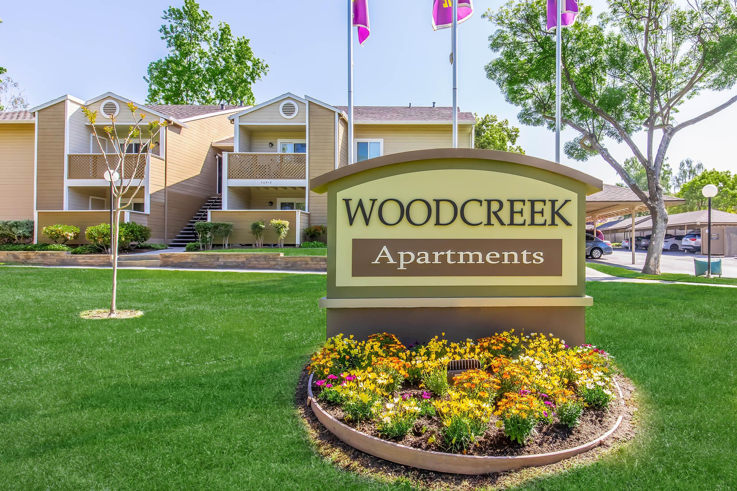 Sign for Woodcreek Apartments in front of a well-maintained building. The sign features colorful flowers and greenery, with several flags above it, indicating a welcoming environment. The complex includes multiple stories and has a nicely landscaped lawn.