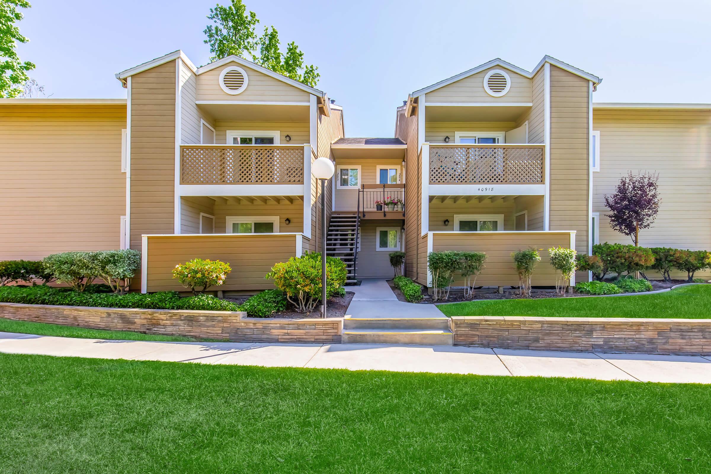 Two-story apartment building with a light brown façade, featuring balconies on the upper level. Well-maintained green lawn and landscaped shrubs in front. A pathway leads to the entrance, with stairs to the upper apartments visible. Clear blue sky above.