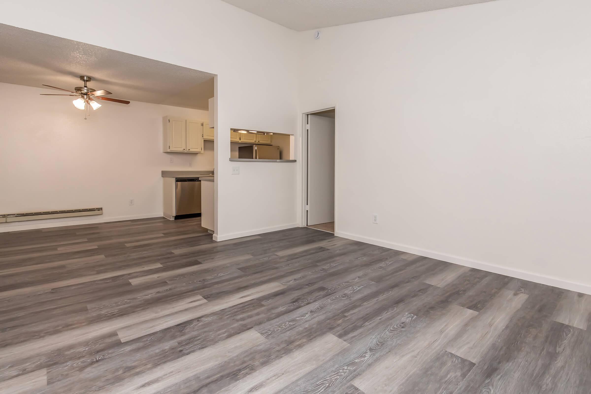A spacious, empty room with light-colored walls and a modern ceiling fan. The floor features laminate planks in gray tones, creating a contemporary look. In the background, there is an open doorway leading to a kitchen area, with a small bar counter visible. Bright natural light fills the space.