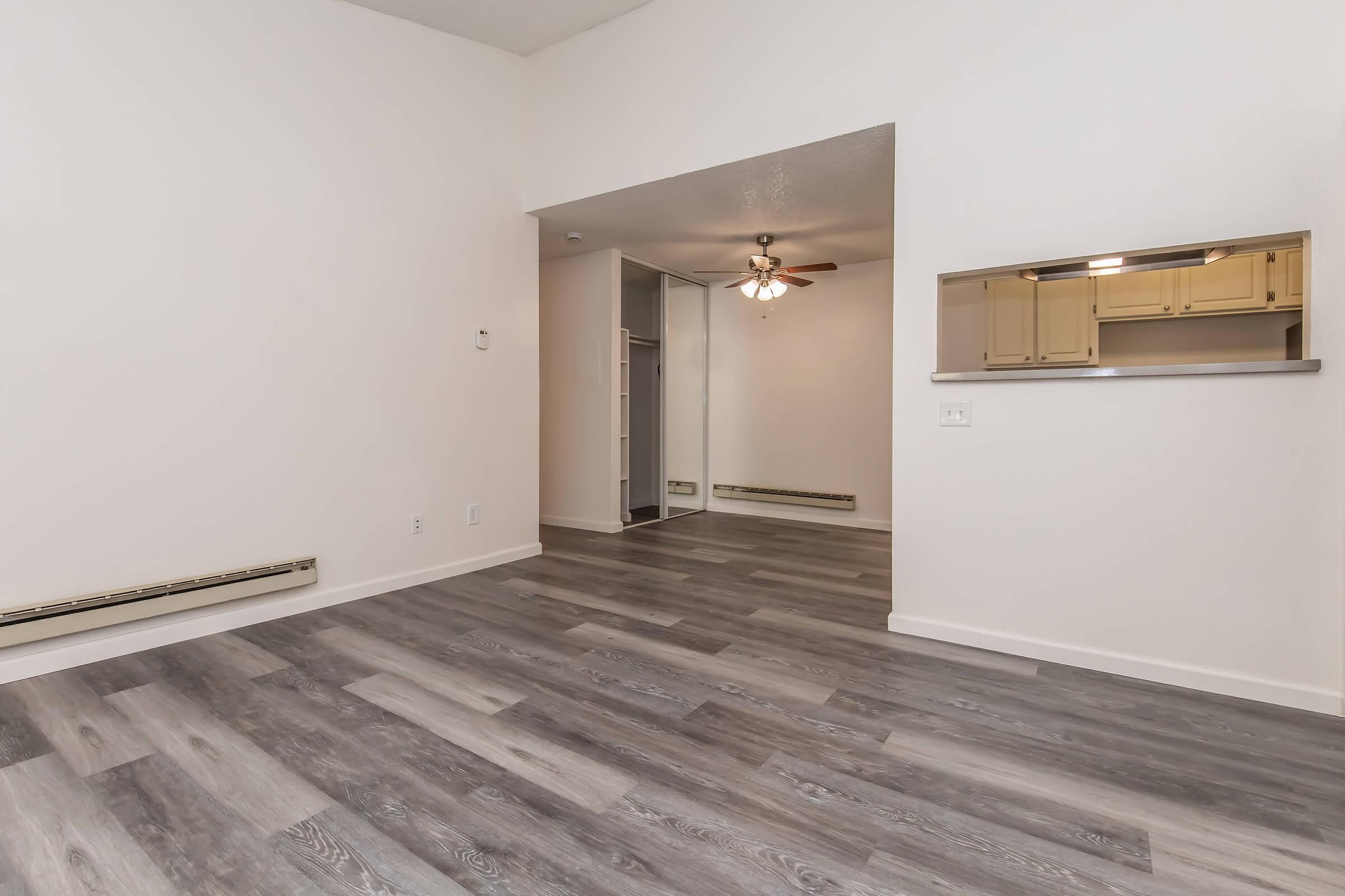 A spacious, empty room featuring light-colored walls and a ceiling fan. The floor is covered with gray laminate, and there is a mirrored wall on one side. A small opening leads to a kitchen area with light-colored cabinets visible in the background. Natural light enhances the open feel of the space.