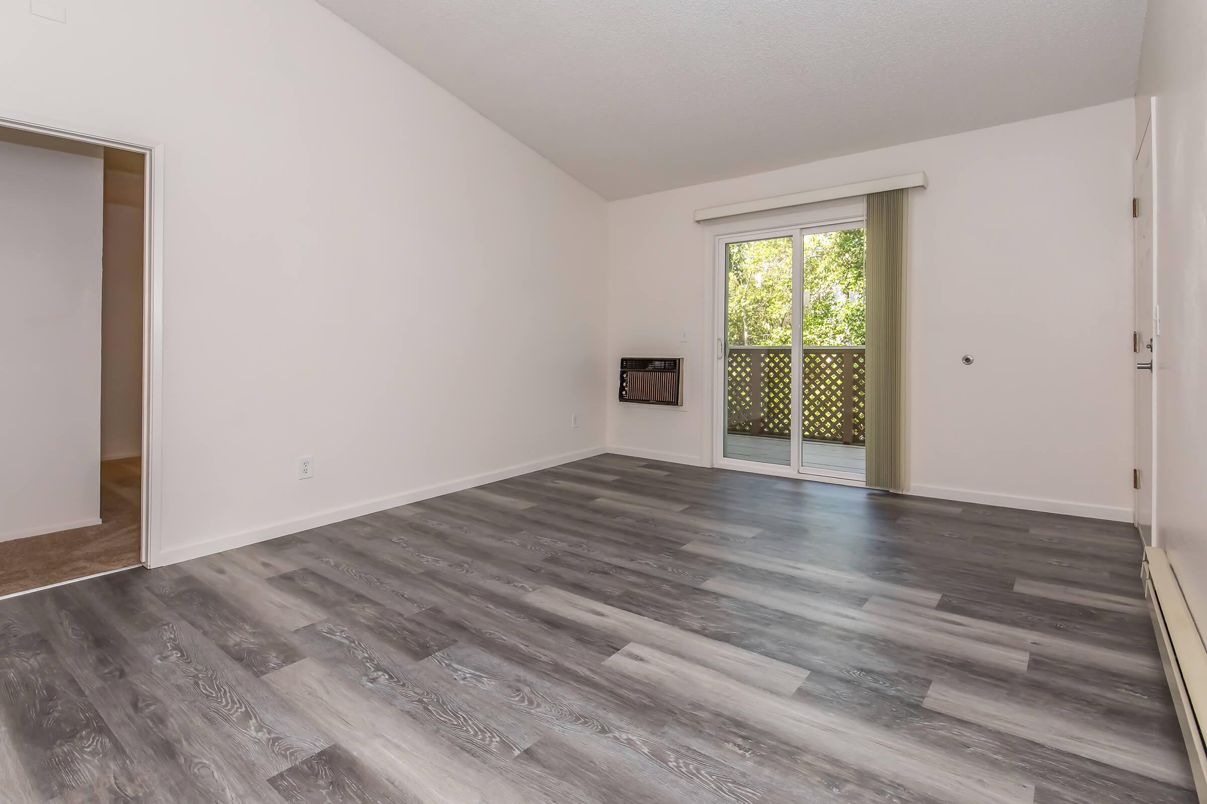 A spacious, empty room with light-colored walls and wood-style flooring. There is a sliding glass door leading to a balcony, allowing natural light to enter. An air conditioning unit is mounted on the wall, and an adjacent doorway leads to another room. The overall atmosphere is bright and airy.
