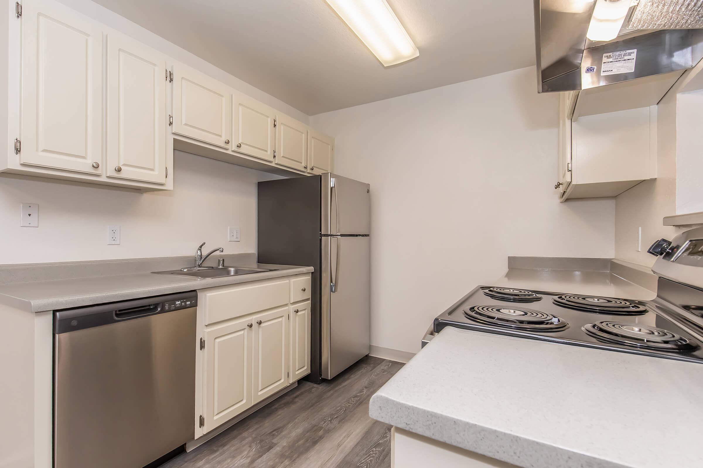 A modern kitchen featuring light-colored cabinets, a stainless steel refrigerator, a dishwasher, and an electric stove. The countertop is sleek and gray, and the space is well-lit with overhead lighting. The flooring is a light wood or laminate finish, creating a clean and open atmosphere.