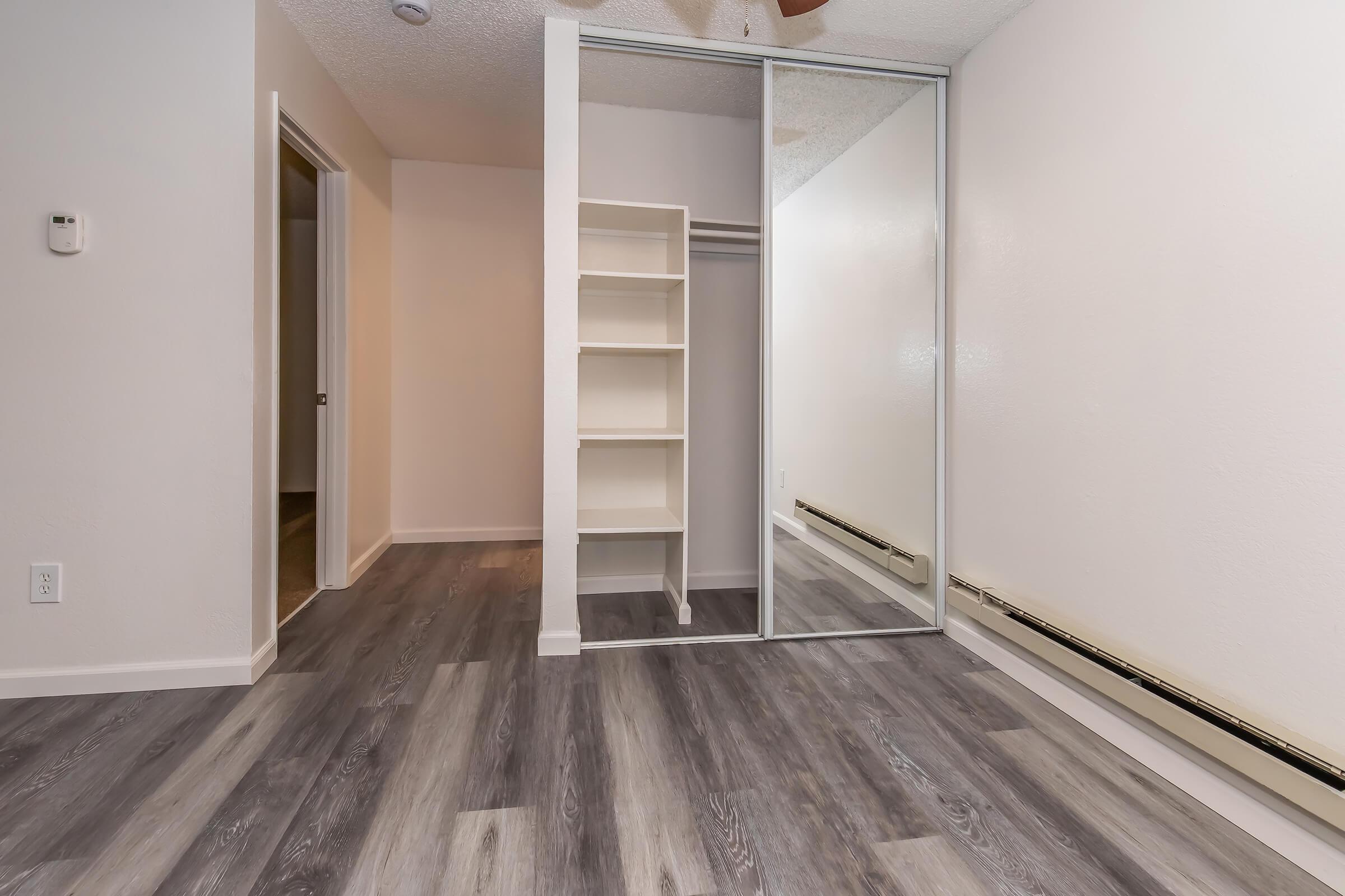 Interior view of a room featuring laminate flooring and a mirrored closet. The space has light-colored walls and a ceiling fan. There is a white shelving unit built into the wall on one side. The room is well-lit, showcasing a clean and modern design with a doorway leading to another room visible in the background.