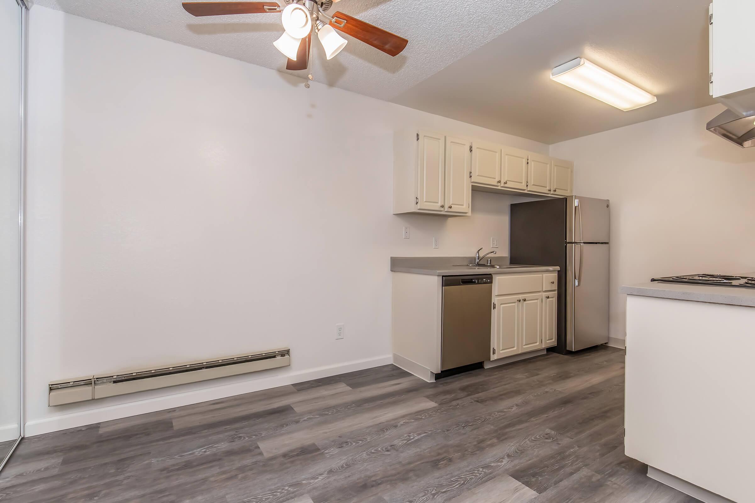 A modern kitchen area featuring light-colored cabinets, a stainless steel refrigerator, and an electric stove. The flooring is a light gray laminate, and there is a ceiling fan with light fixtures. The walls are painted white, creating a bright and airy atmosphere.