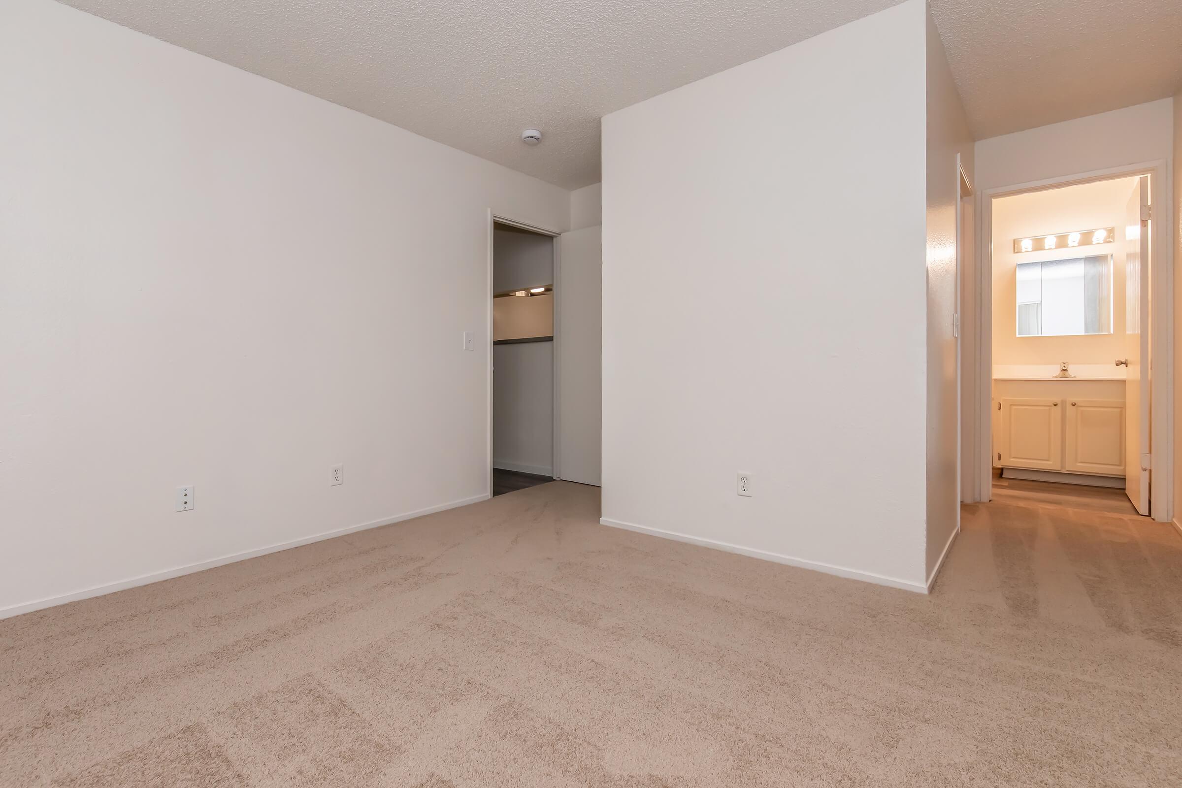A spacious, empty room with light beige carpet and white textured walls. A doorway on the left leads to a closet, and another door on the right opens to a bathroom with bright lighting. Natural light filters in, highlighting the clean, simple design of the space.