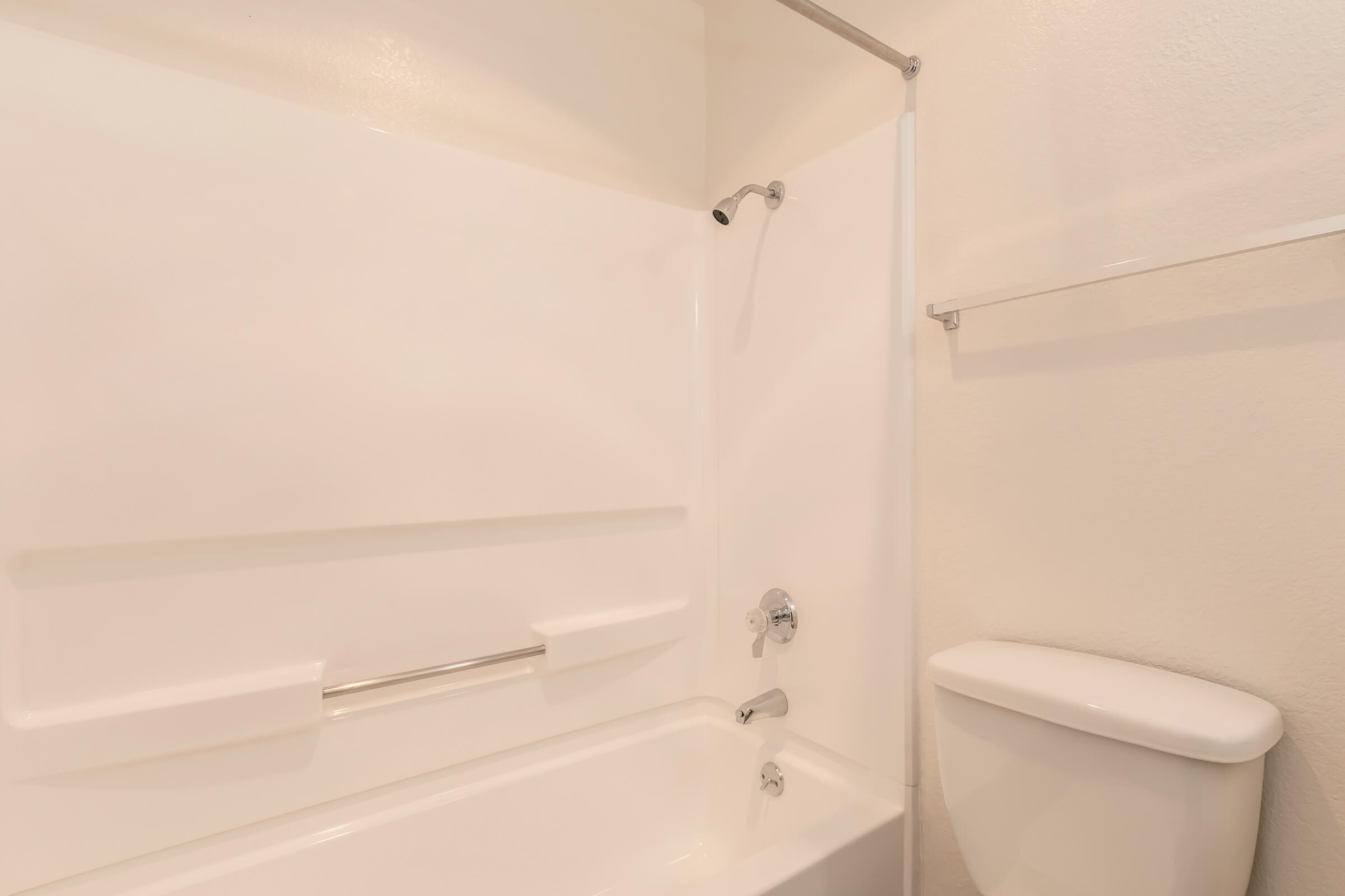 A clean and simple bathroom setup featuring a white bathtub with a showerhead, a wall-mounted shelf, and a white toilet. The walls are painted a light color, creating a bright and minimalistic atmosphere.