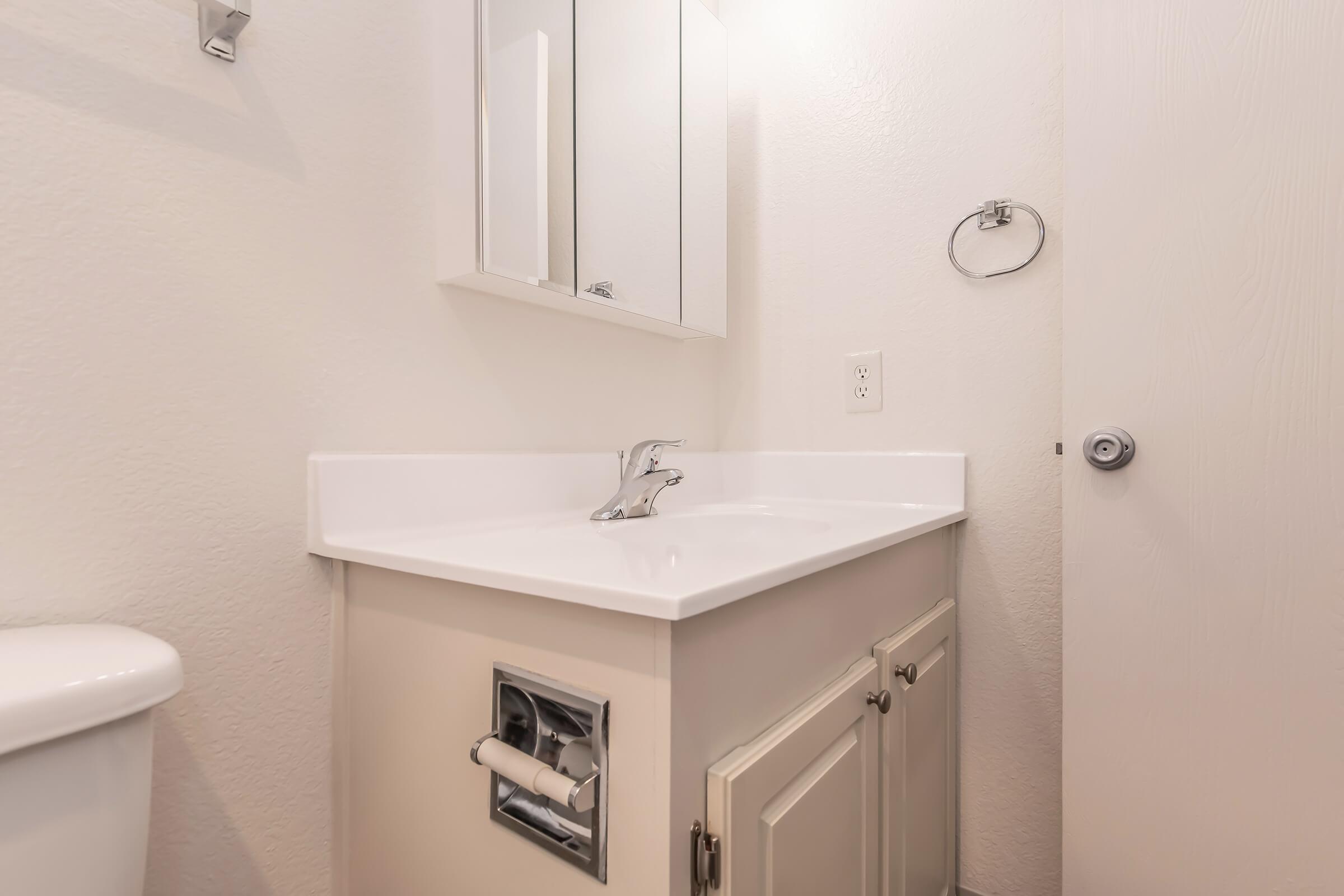 A modern bathroom featuring a white sink with a silver faucet, a small cabinet underneath, a mirror above the sink, and a towel holder on the wall. The walls are light-colored, and there is a door leading out of the room. A toilet is visible to the left.