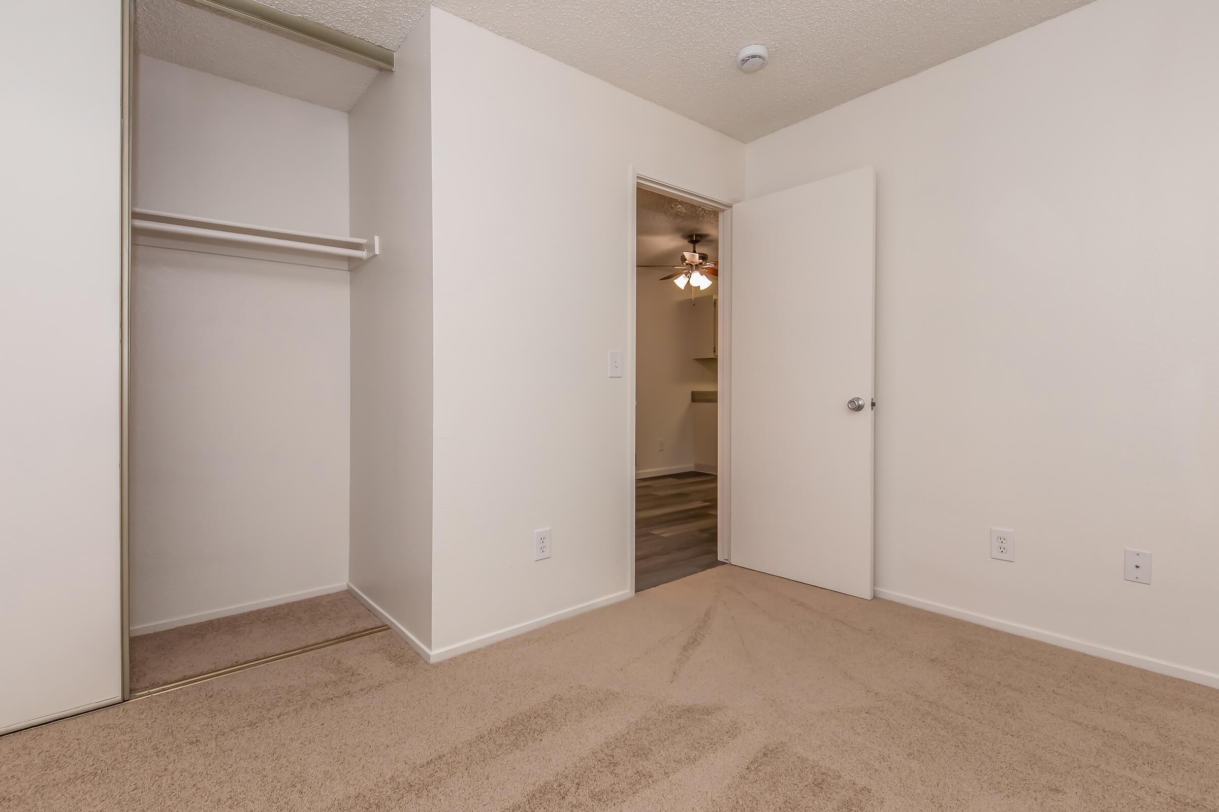 Interior view of a small, light-colored room featuring a carpeted floor, a closet with sliding doors on the left, and an open door leading to another room. The space is bright and minimalistic, with a neutral color palette and a ceiling fan visible in the background.