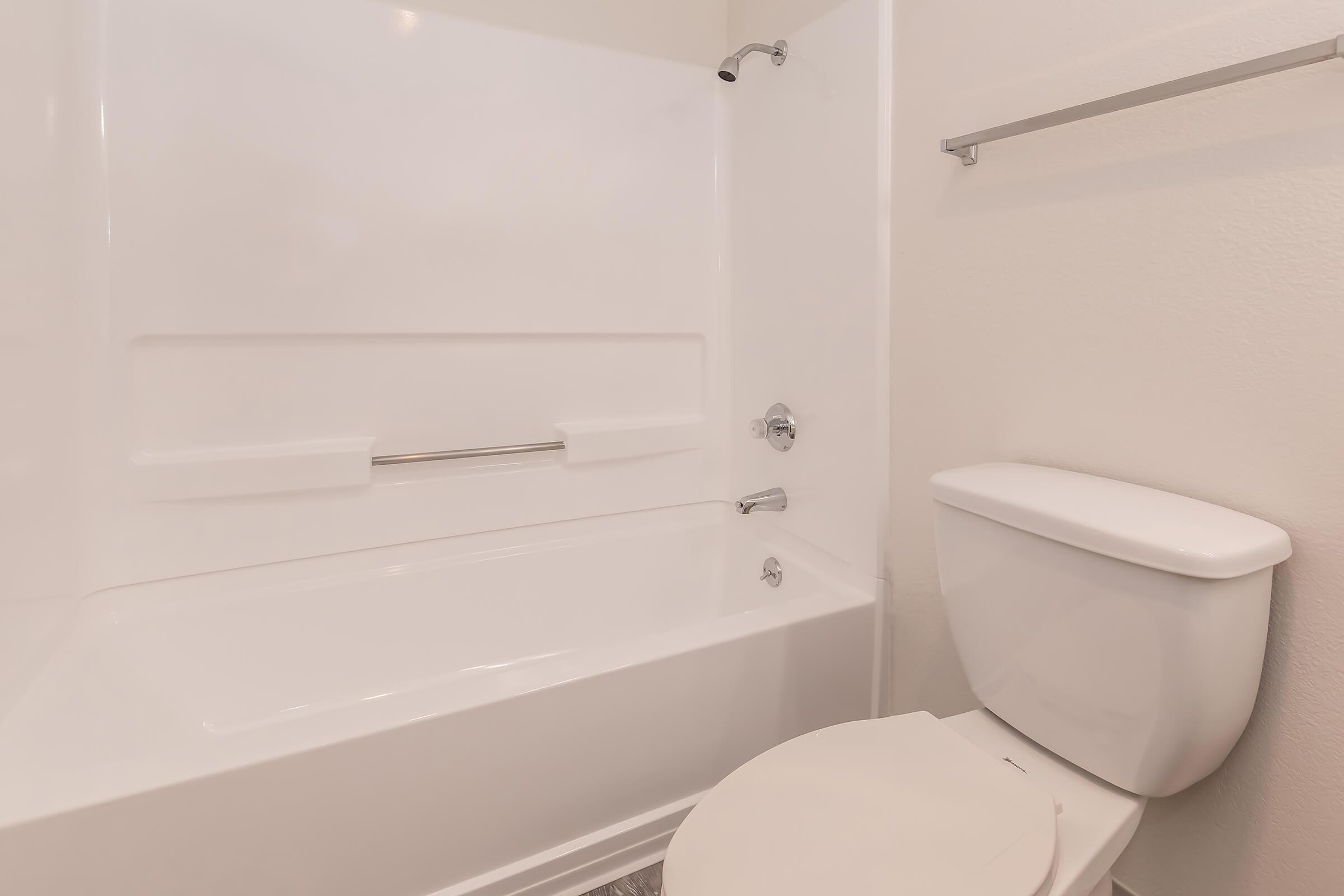 A clean and modern bathroom featuring a white bathtub with a showerhead, a simple toilet, and a towel bar on the wall. The walls are light-colored, providing a bright and spacious feel to the room.