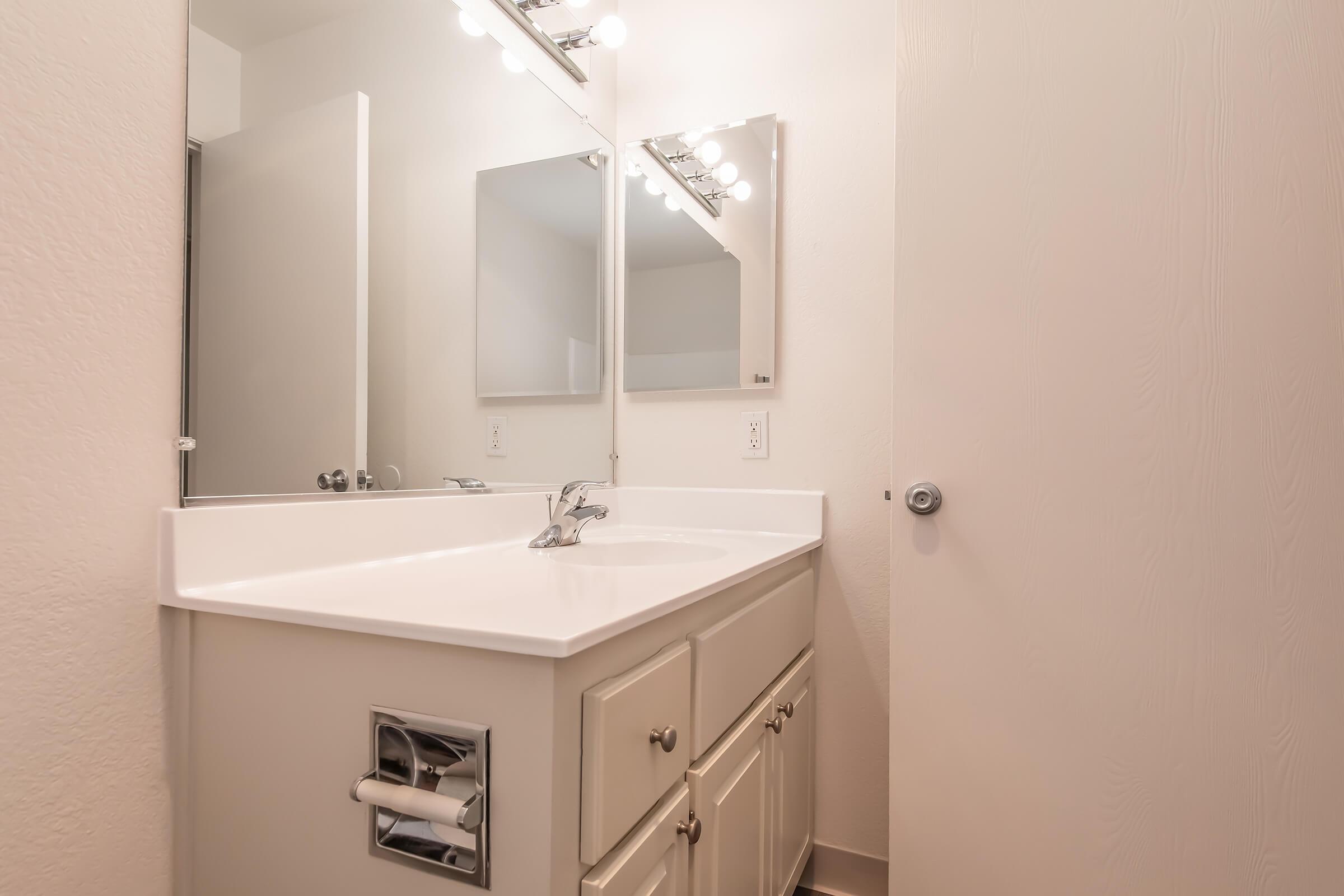 A clean, modern bathroom featuring a white countertop with a sink, two mirrors above, and light fixtures overhead. The room has light-colored walls and a door to the right. A toilet paper holder is visible beneath the countertop. The space is well-lit and looking tidy.
