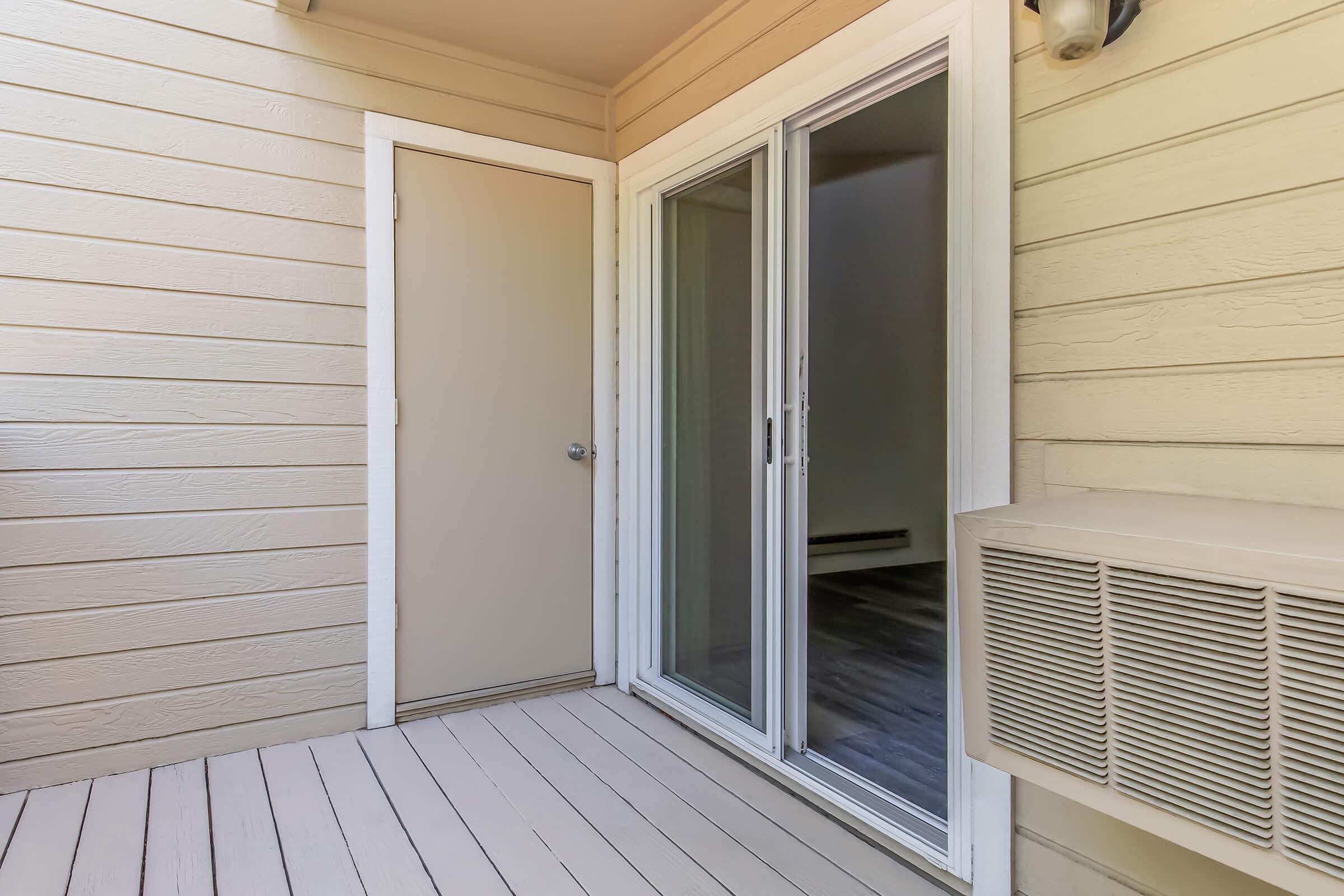 A view of a small outdoor patio area featuring a sliding glass door and a closed door. The floor is made of wooden planks, and there is an air conditioning unit mounted on the wall. The walls are made of light-colored wood siding. The interior of the room beyond the glass door is not visible.