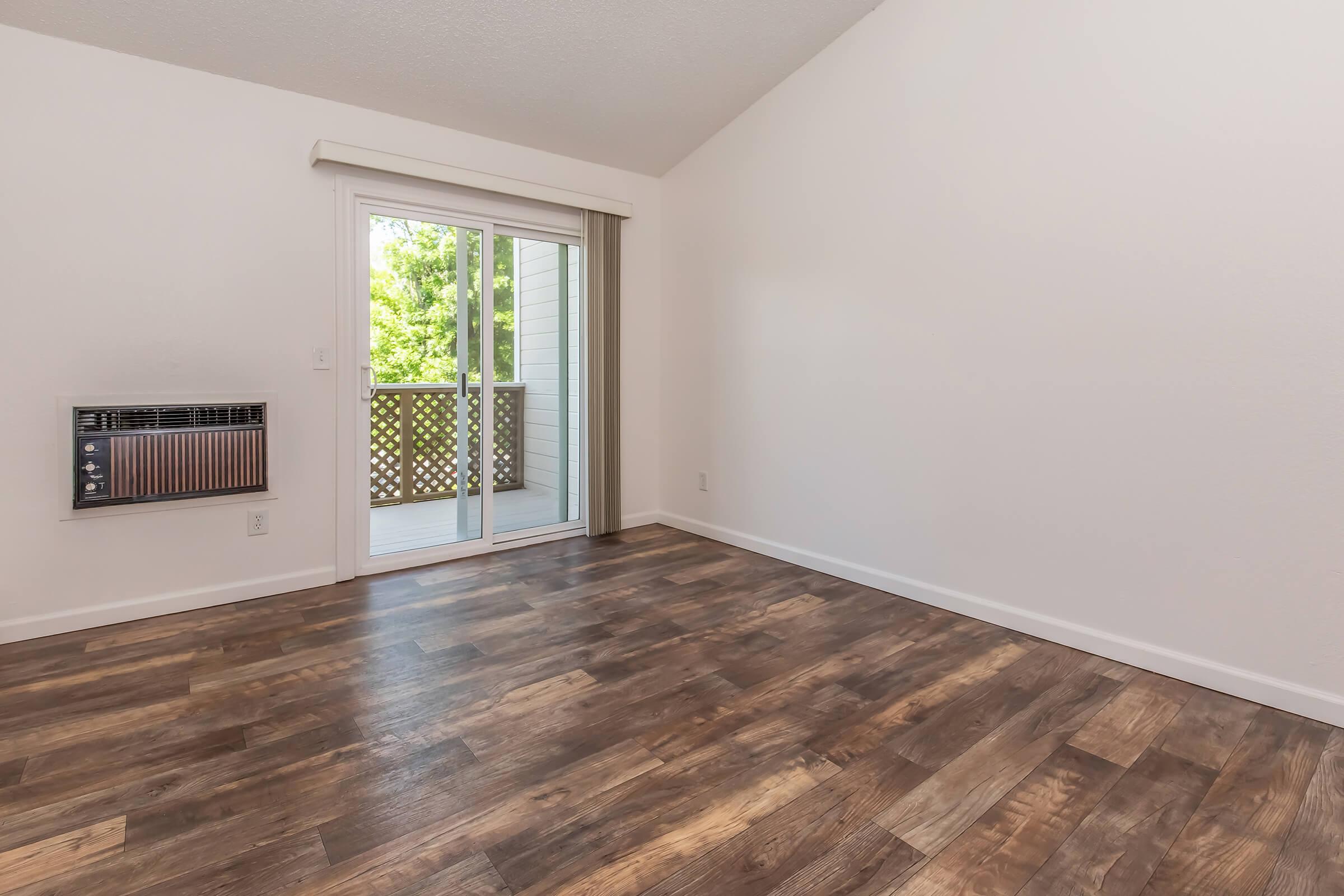 Empty room with hardwood floors, featuring a large sliding glass door leading to a small balcony. Walls are painted white, and there is a wall-mounted air conditioning unit on the left. The space is well-lit with natural light coming in through the door.