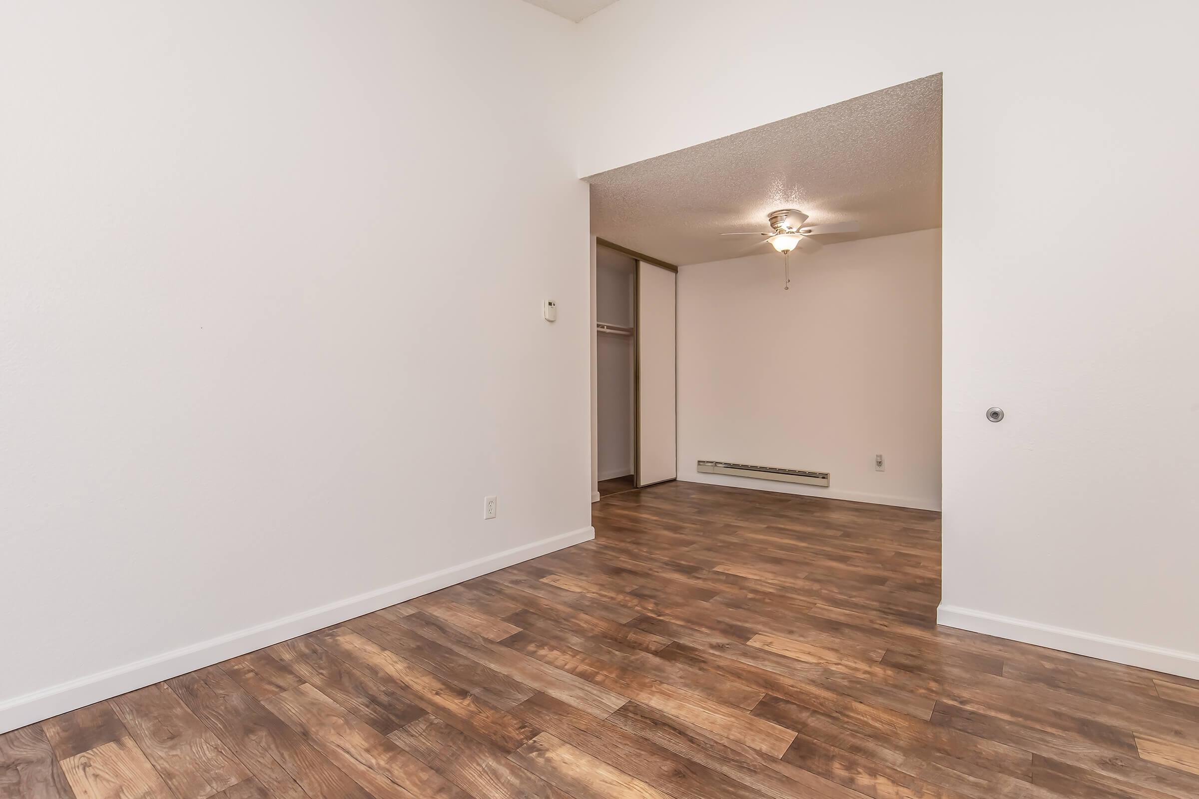 Interior view of a spacious room with light-colored walls and wooden flooring. A ceiling fan is visible in the background, along with an open doorway leading into another room. The space is bright and uncluttered, creating a welcoming atmosphere.