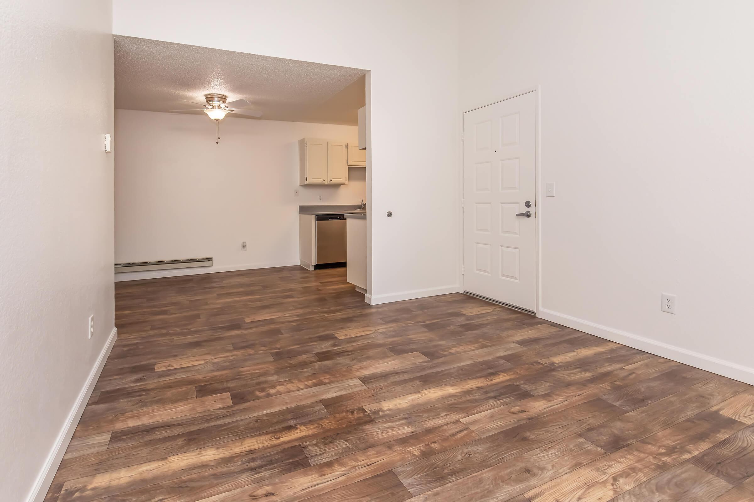 A spacious, empty living area with light-colored walls and hardwood floors. A ceiling fan is visible, and there is an open archway leading to a kitchen area in the background. A door on the right leads to an outer area. The room is well-lit, creating a welcoming atmosphere.