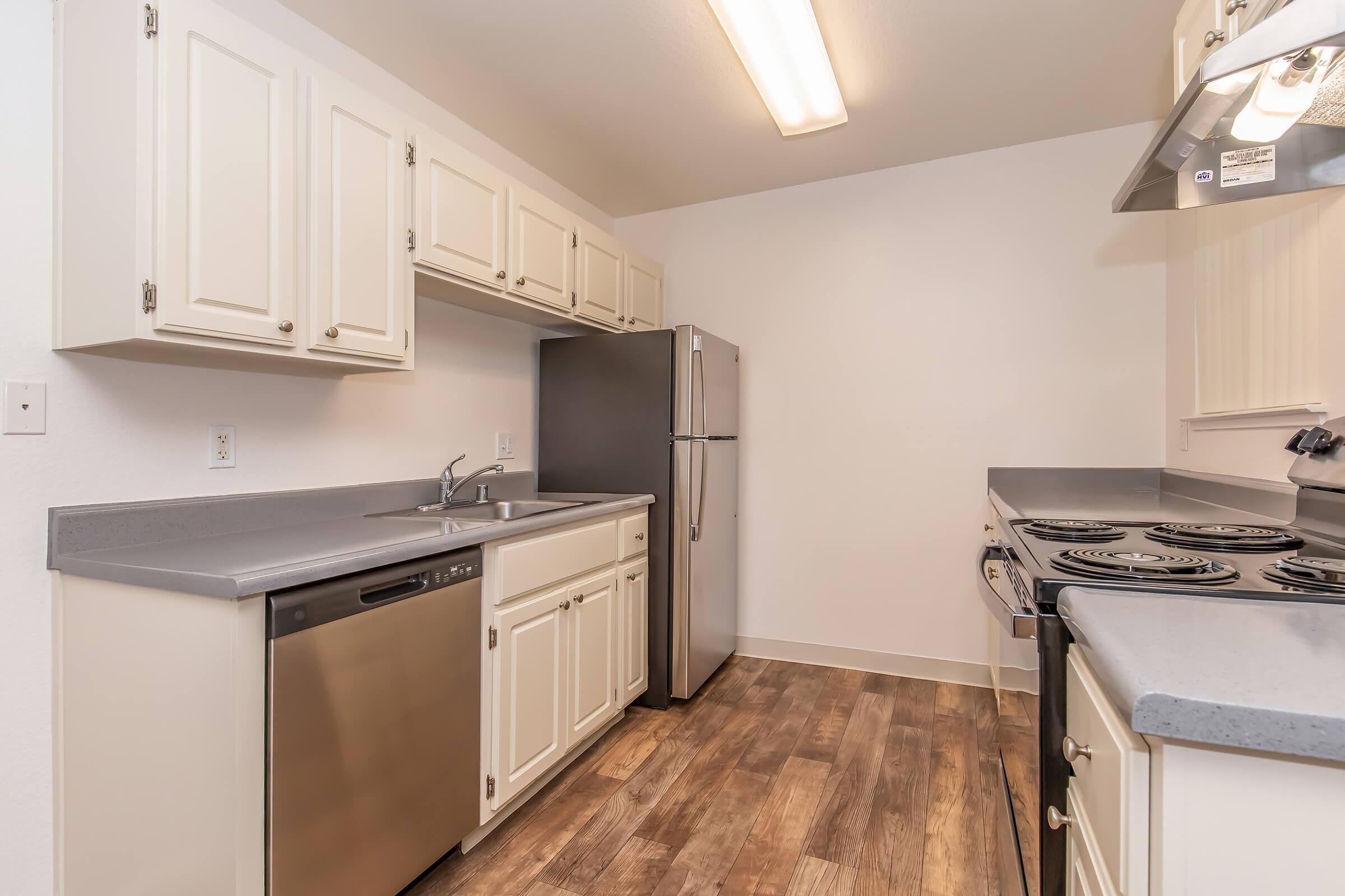 A modern kitchen featuring white cabinetry, stainless steel appliances including a refrigerator and dishwasher, and a grey countertop. The space has a neutral wall color and wood-like flooring, providing a clean and bright ambiance. The kitchen includes a stove and ample storage.