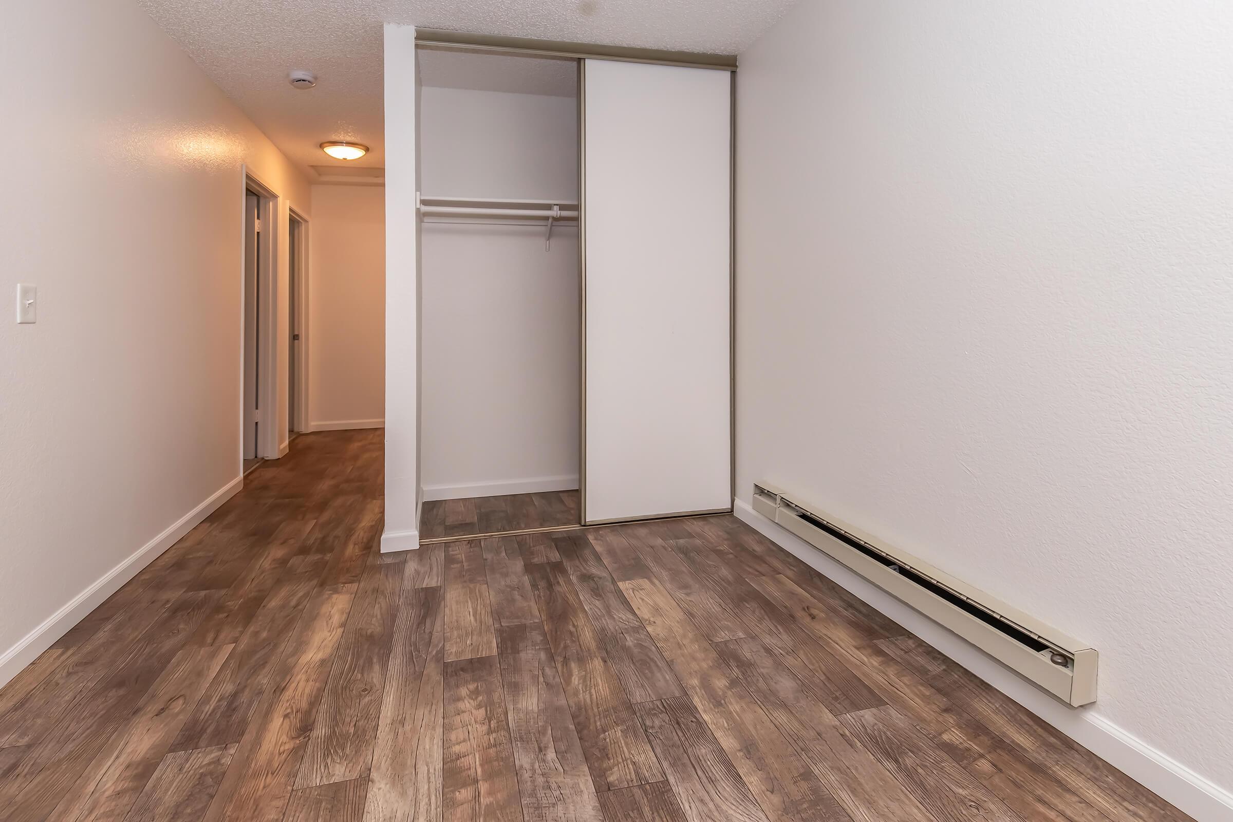 Empty room with light-colored walls and wood-like flooring. A sliding closet door is open, revealing an empty closet space. A hallway is visible in the background leading to additional rooms. Baseboard heating is installed along one wall. The overall atmosphere is clean and modern.