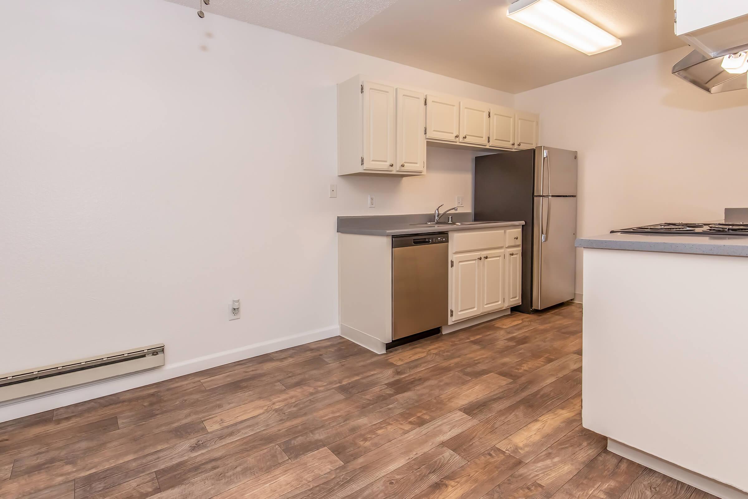 Modern kitchen featuring white cabinets, stainless steel appliances including a refrigerator and dishwasher, and a flat countertop. The space has light-colored walls and wood-like laminate flooring. A window and overhead light provide natural and artificial illumination.